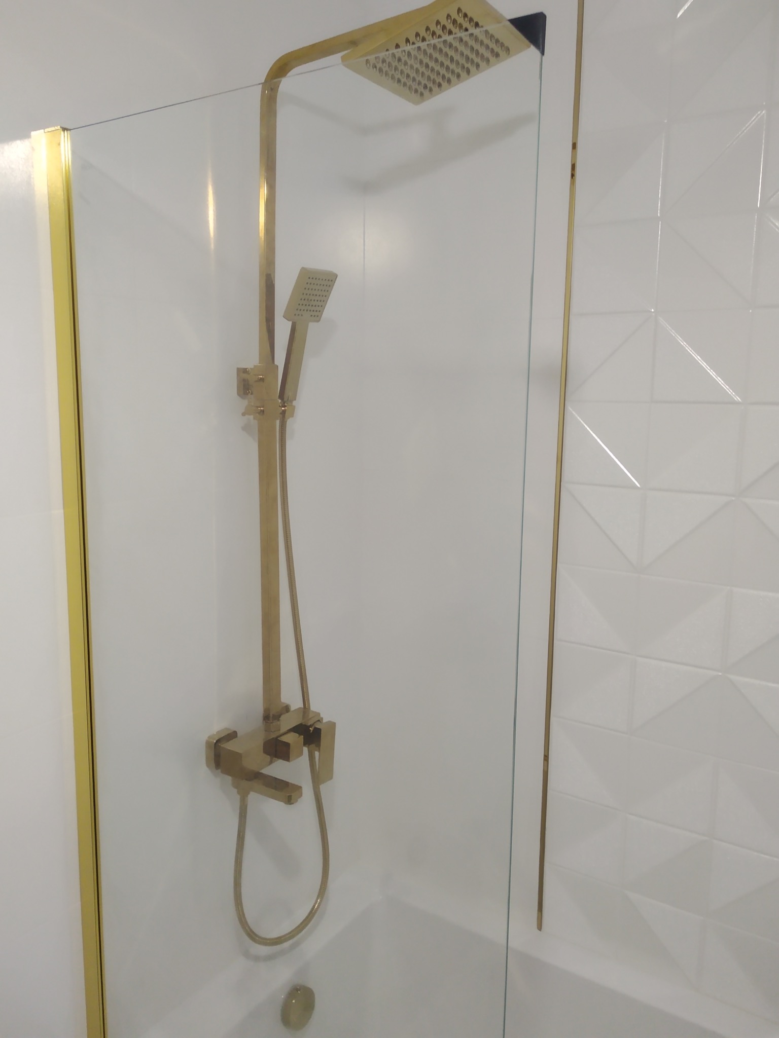 Kabina prysznicowa z złotym wykończeniem, bateria i deszczownica w kolorze złota, białe płytki 3D na ścianie.