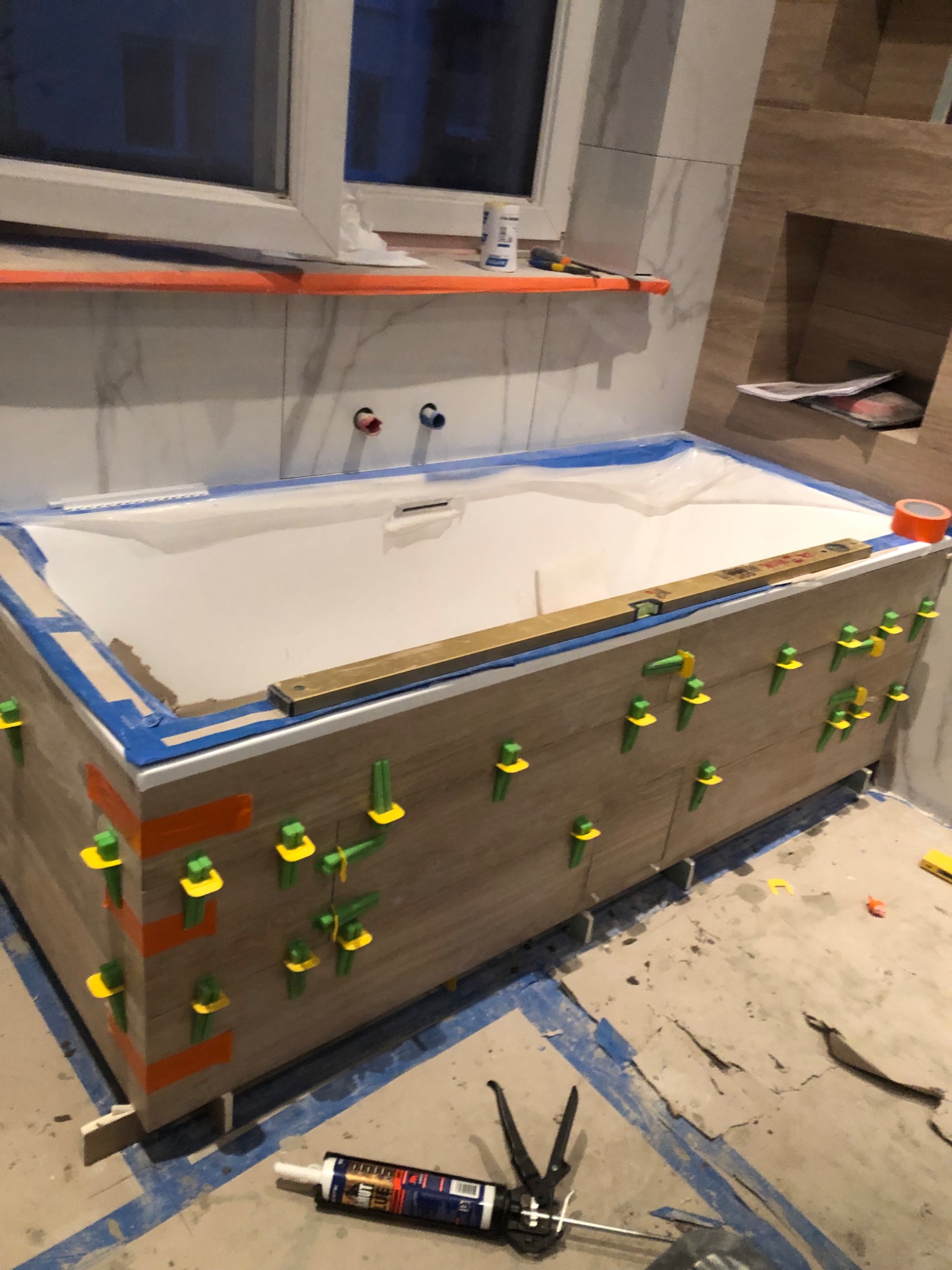 Obudowa wanny z drewnopodobnych płytek w trakcie montażu, z użyciem poziomicy i klipsów poziomujących, widoczny silikon i narzędzia.