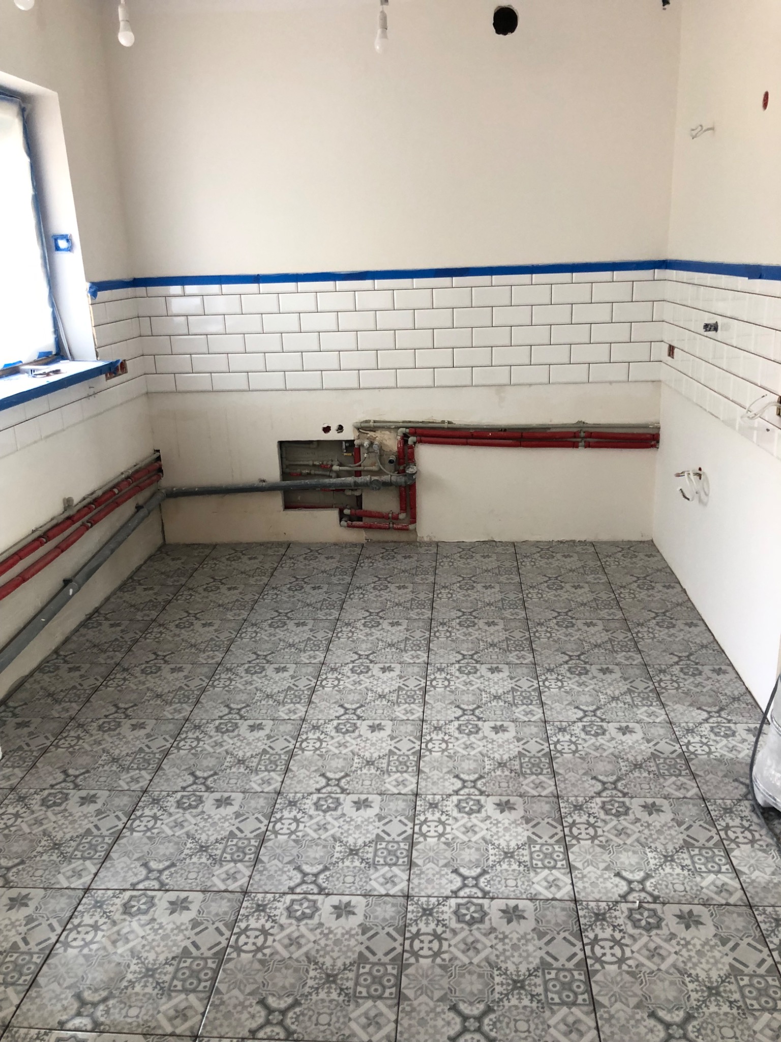 Wnętrze w trakcie prac: położone płytki podłogowe w szaro-białe wzory, częściowo wykończone ściany z białymi, prostokątnymi płytkami, zabezpieczone niebieską taśmą malarską, widoczne instalacje...