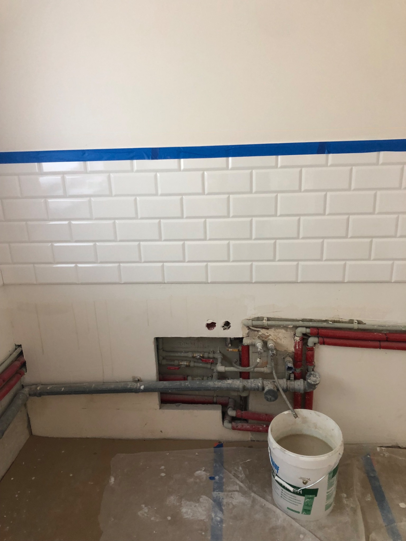 Białe płytki metra na ścianie łazienki z niebieską taśmą malarską, odsłonięte rury wodne i kanalizacyjne w ścianie, wiadro z zaprawą na podłodze, etap remontu.
