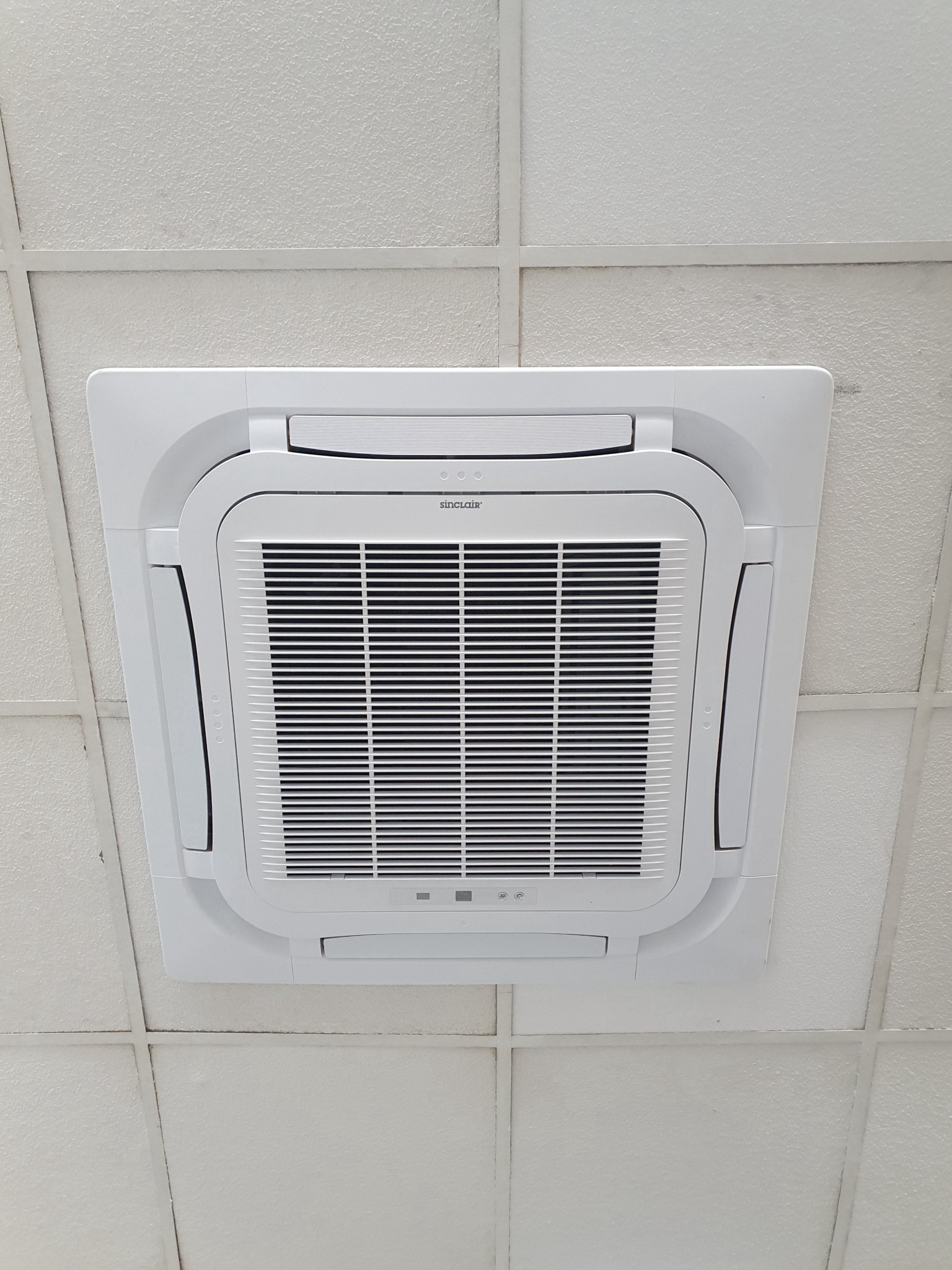 Sufitowy klimatyzator kasetonowy marki Sinclair, zamontowany w biurze, widoczny od dołu na tle białego sufitu kasetonowego.