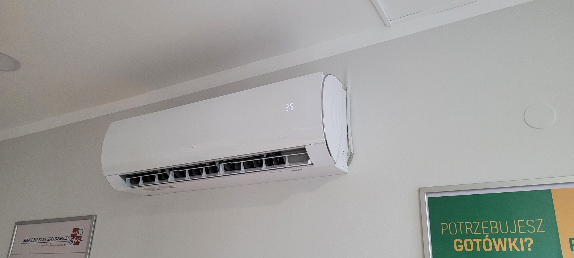 Biała jednostka klimatyzacji ściennej zamontowana w jasnym pomieszczeniu biurowym, widoczny wyświetlacz z temperaturą 25 stopni Celsjusza, w tle plakat reklamowy banku.