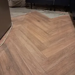 Układanie parkietu w jodełkę z płytek imitujących drewno w trakcie remontu kuchni, widoczne niedociągnięcia przy ścianie i pod szafkami.