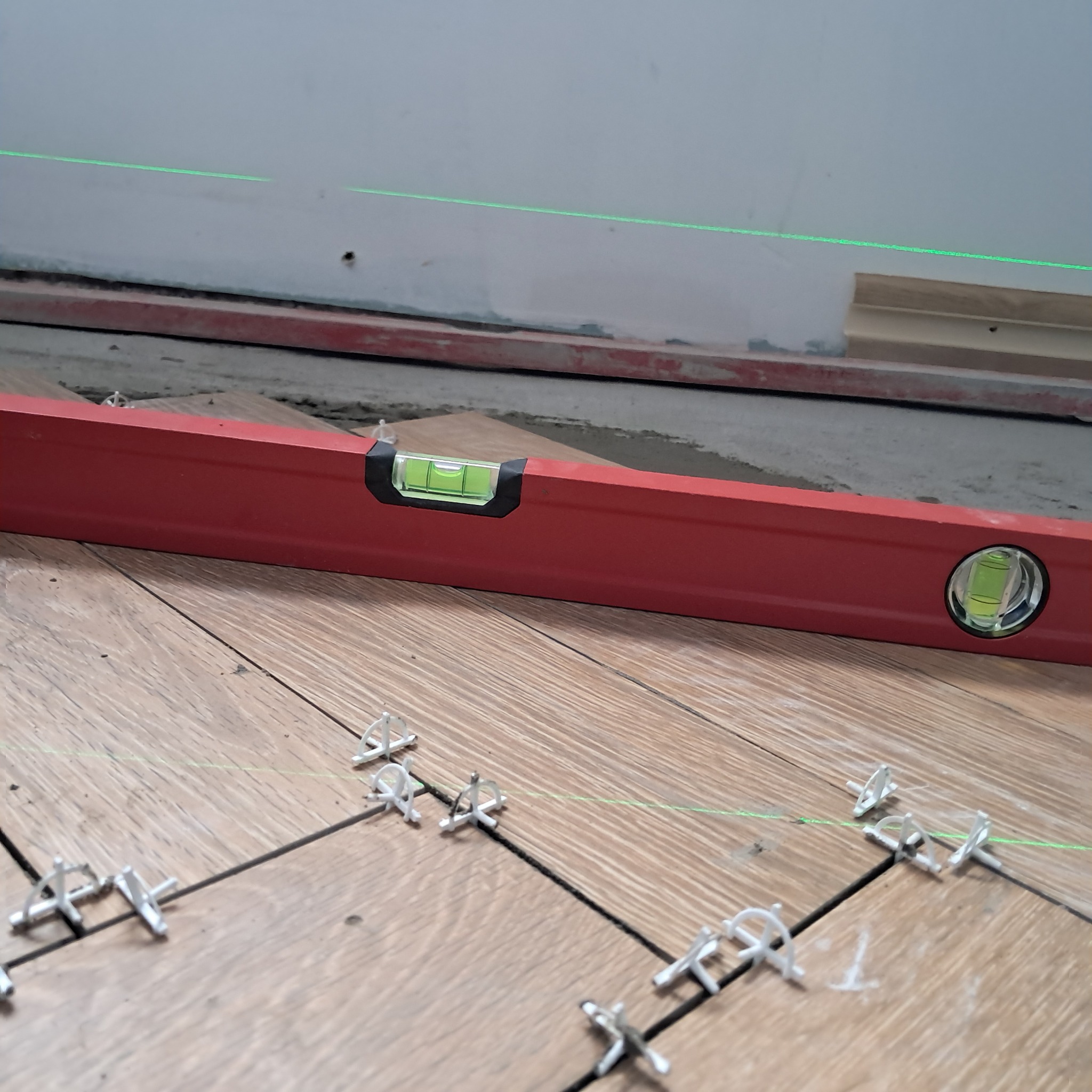 Poziomica laserowa i tradycyjna na ułożonych płytkach imitujących drewno, z krzyżykami dystansowymi w fugach.