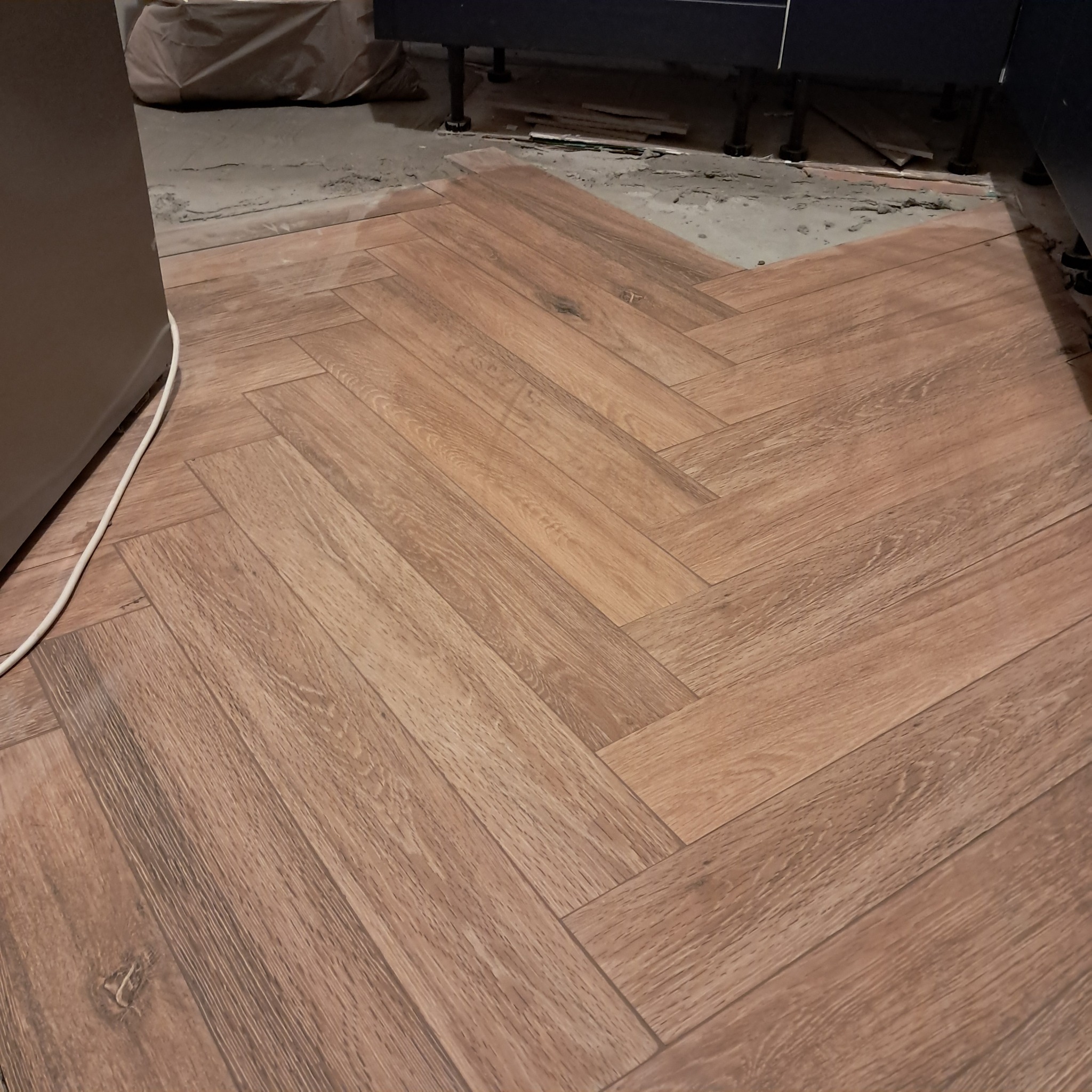 Układanie parkietu w jodełkę z płytek imitujących drewno w trakcie remontu kuchni, widoczne niedociągnięcia przy ścianie i pod szafkami.