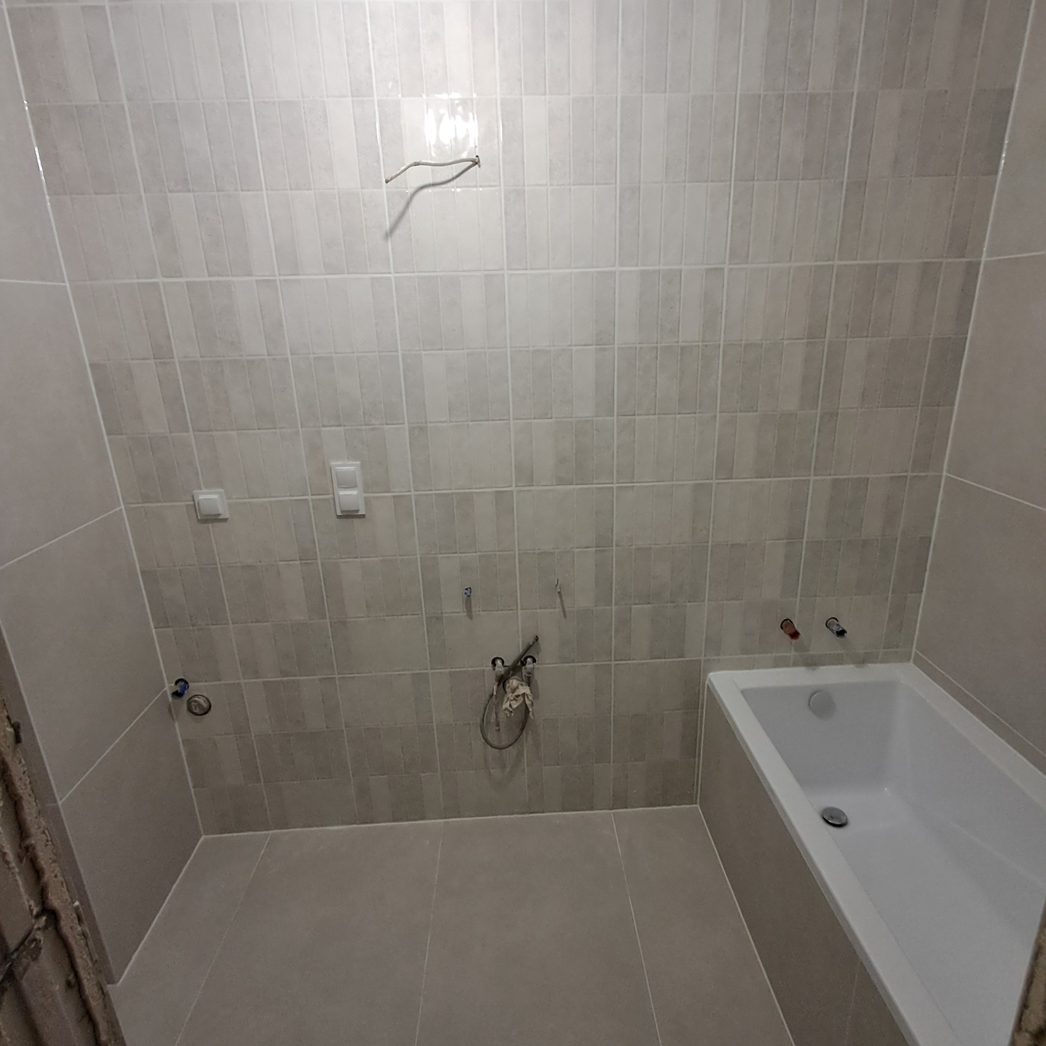 Ściany łazienki wyłożone szarymi, pionowymi płytkami, z widocznymi instalacjami wodnymi, gniazdkami elektrycznymi i częściowo zamontowaną białą wanną.