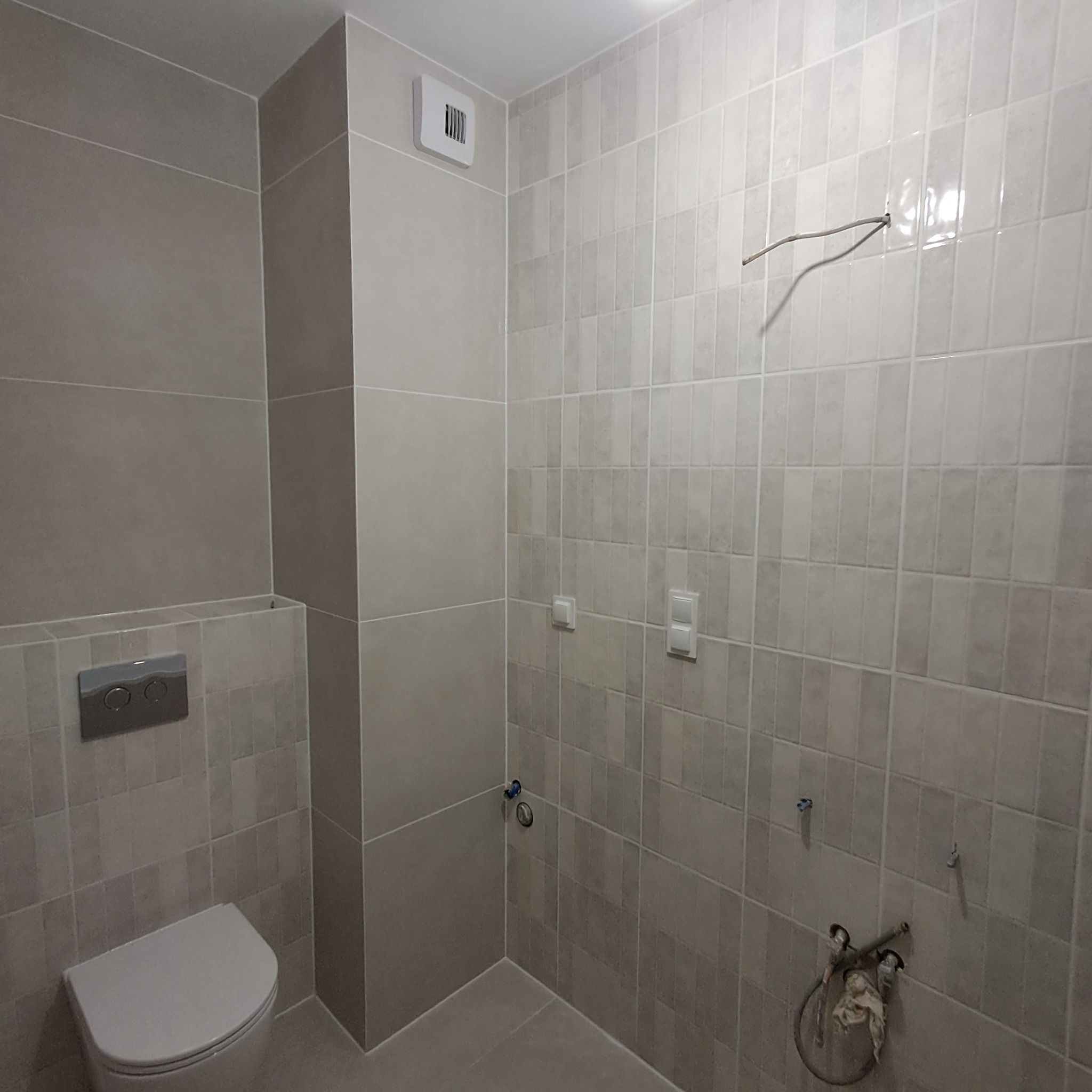 Szare, pionowo ułożone płytki w trakcie remontu łazienki, widoczne wystające kable, niezamaskowane przyłącza wodne oraz częściowo zamontowana toaleta podtynkowa.