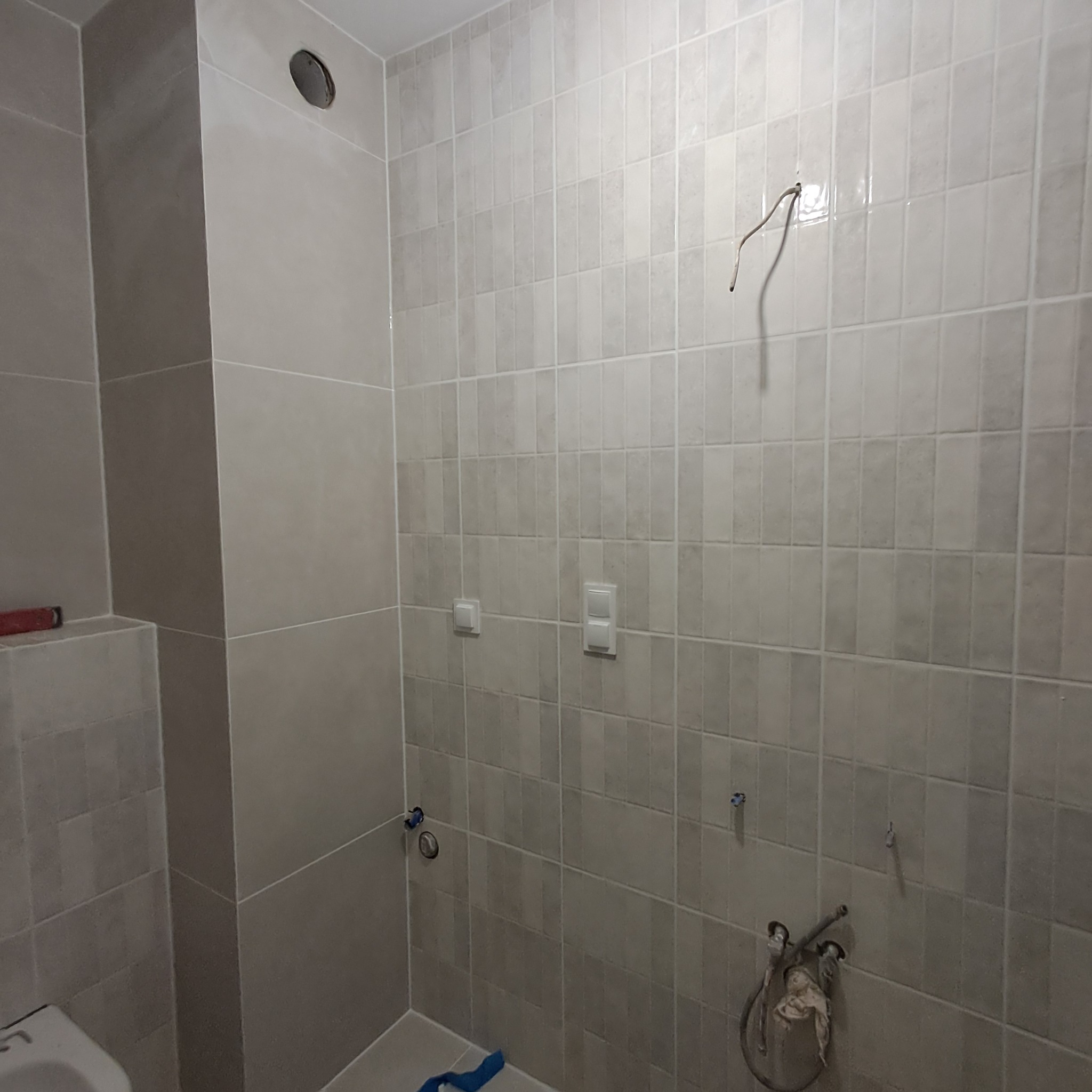 Ściana łazienki wyłożona szarymi, pionowymi płytkami ceramicznymi, widoczne wystające kable, instalacja wodna i otwór na oświetlenie sufitowe, etap prac wykończeniowych.