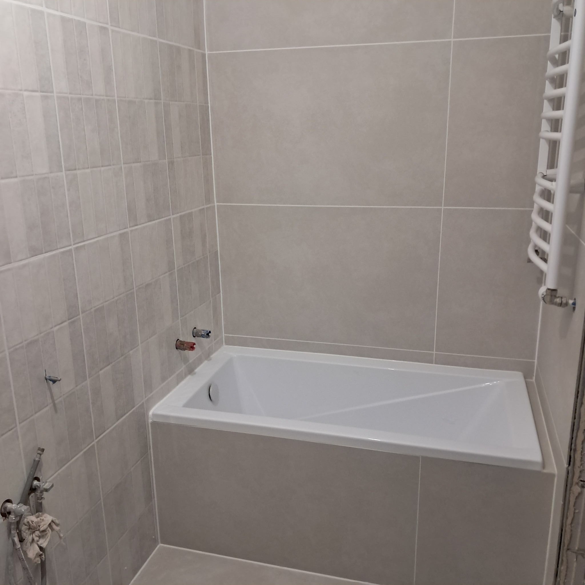 Wykończona łazienka z wanną obudowaną szarymi płytkami, widoczne przyłącza wodne i grzejnik drabinkowy, szare płytki na ścianach ułożone w dwóch różnych formatach.