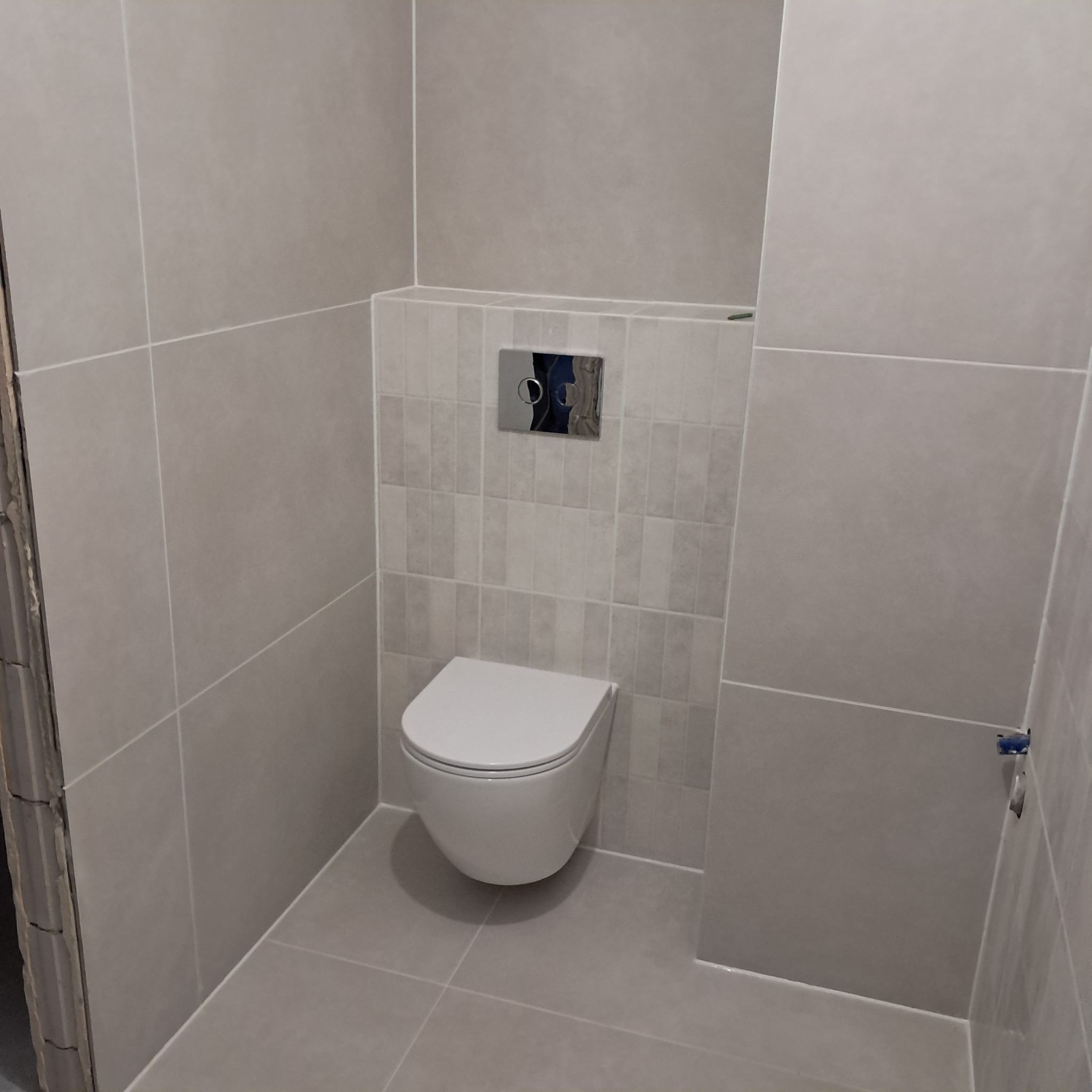 Nowoczesna, podwieszana toaleta w łazience wykończonej szarymi, prostokątnymi płytkami z dekoracyjnym pasem pod chromowanym przyciskiem spłuczki.