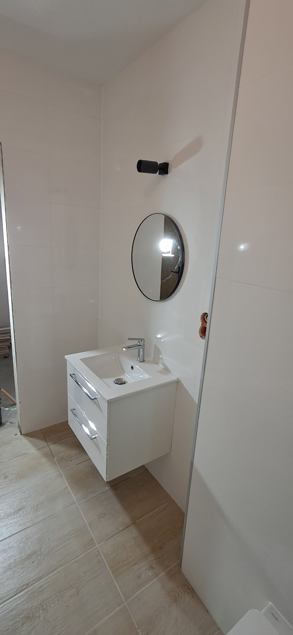 Nowoczesna łazienka z białą umywalką nablatową z szafką, okrągłym lustrem w czarnej ramie i minimalistycznym oświetleniem punktowym na białych płytkach ściennych, widoczna podłoga z płytek...