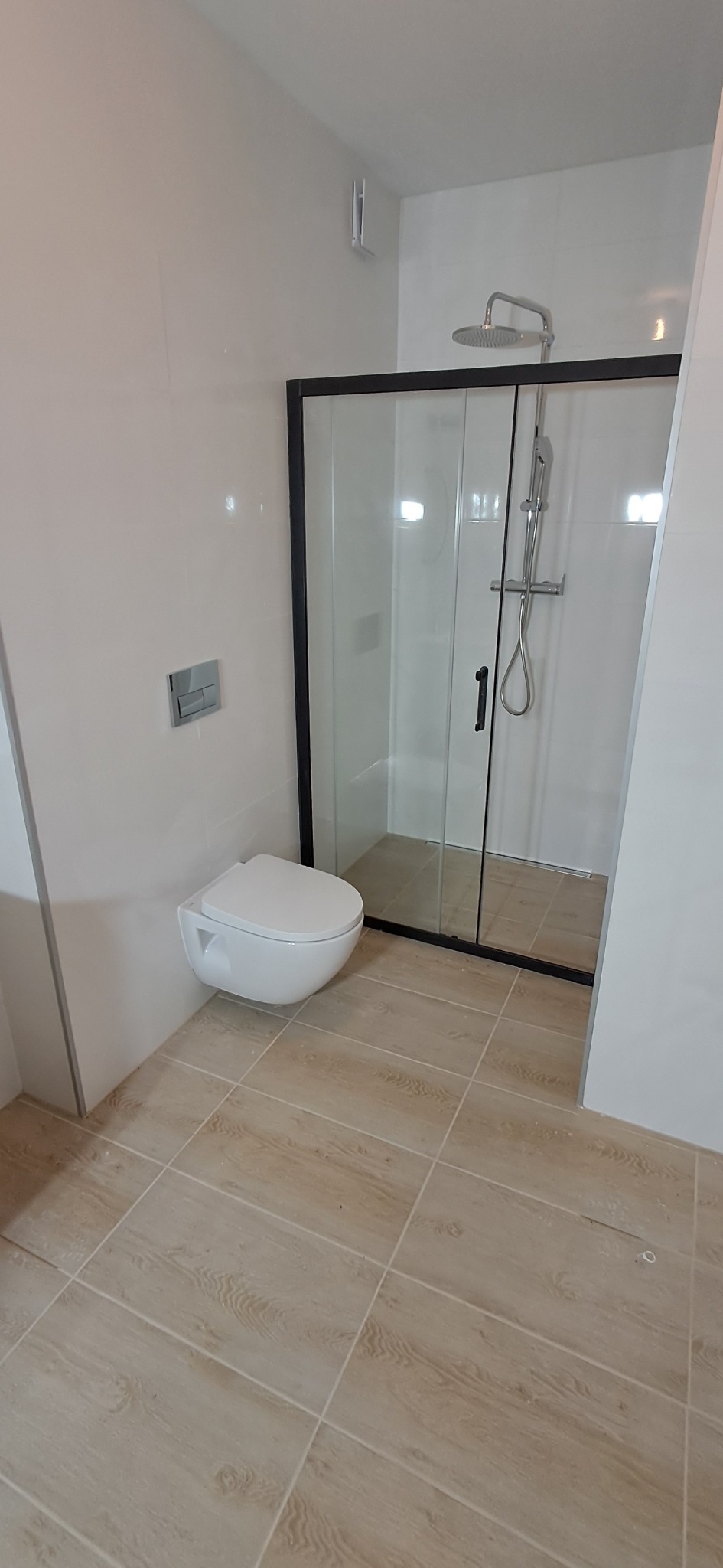 Nowoczesna łazienka z kabiną prysznicową typu walk-in z czarną ramą, wiszącą miską WC i beżowymi płytkami imitującymi drewno na podłodze.