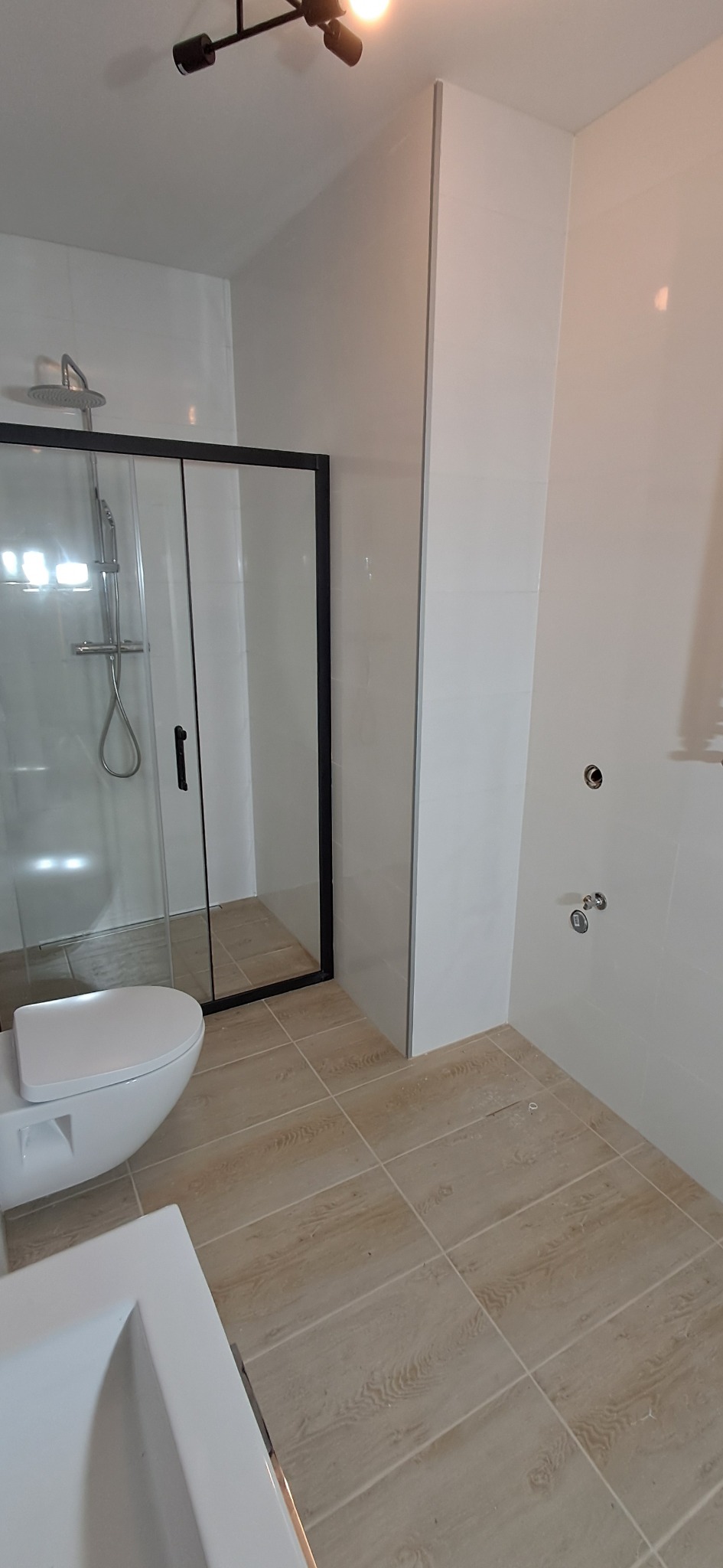 Nowoczesna łazienka z kabiną prysznicową z czarną ramą, podwieszaną toaletą i umywalką, wykończona jasnymi płytkami na ścianach i podłodze imitującymi drewno.