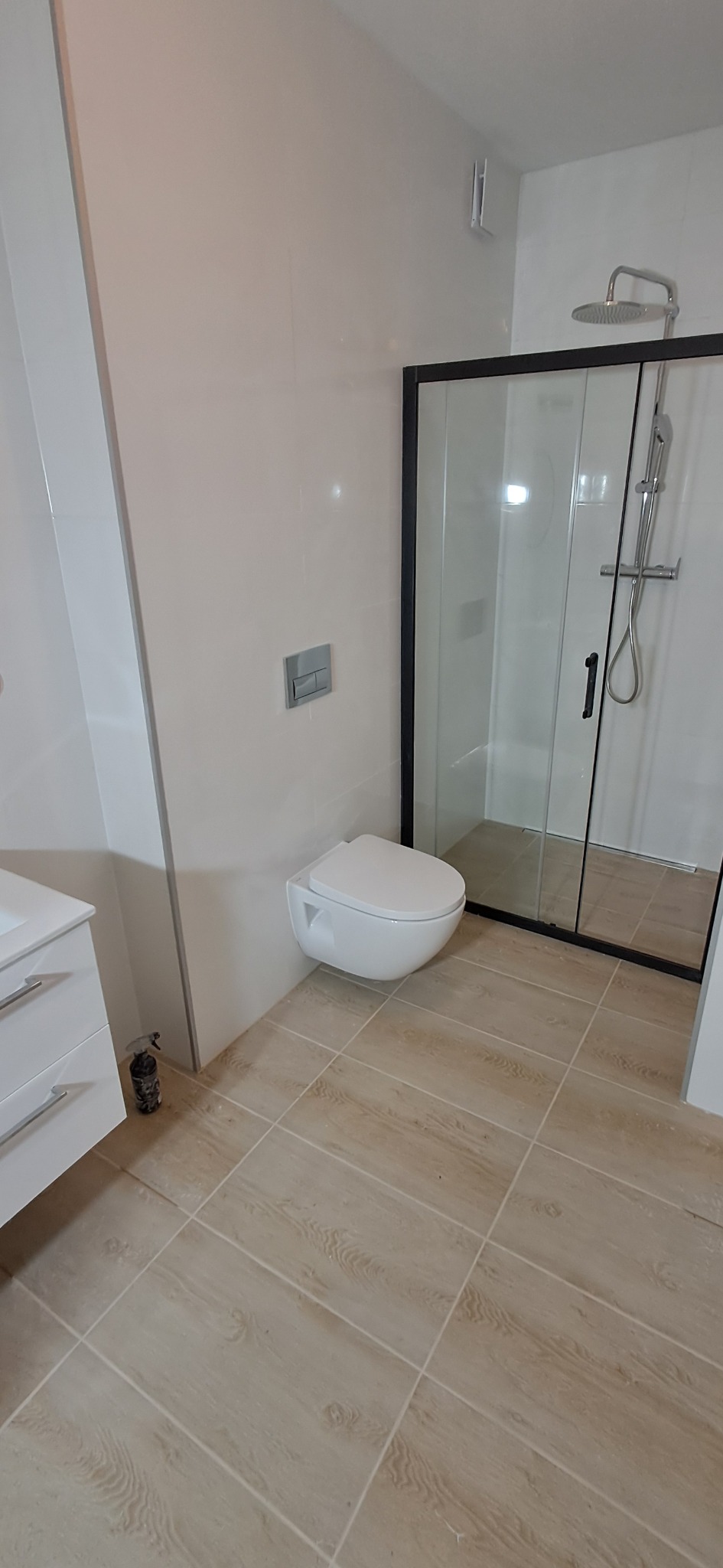 Nowoczesna łazienka po remoncie: wisząca toaleta z przyciskiem spłukującym na ścianie, kabina prysznicowa z czarną ramą, umywalka z szafką i beżowe płytki podłogowe imitujące drewno.