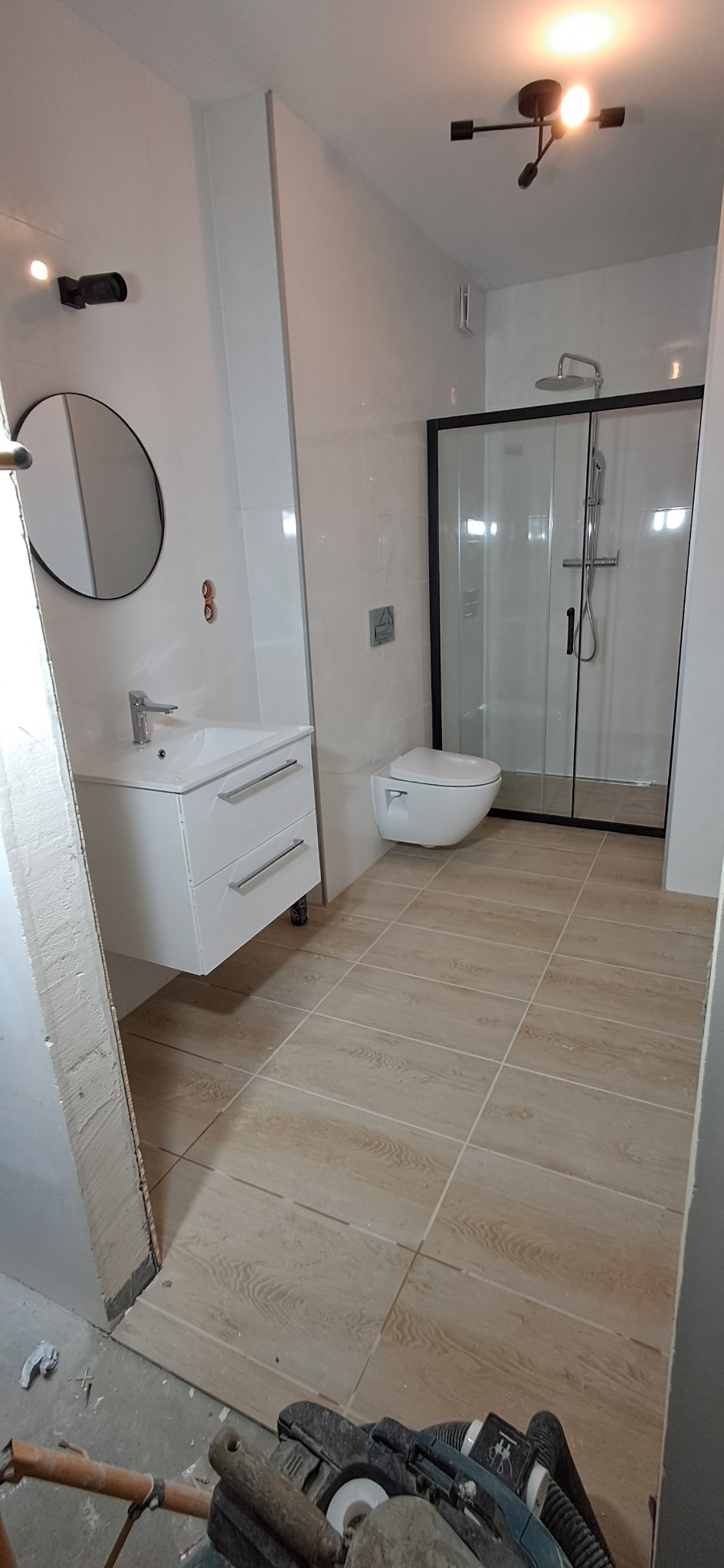 Nowoczesna łazienka z kabiną prysznicową typu walk-in, wiszącą toaletą i umywalką z szafką, wykończona płytkami imitującymi drewno, z widocznymi śladami prac remontowych i narzędziem szlifierskim...