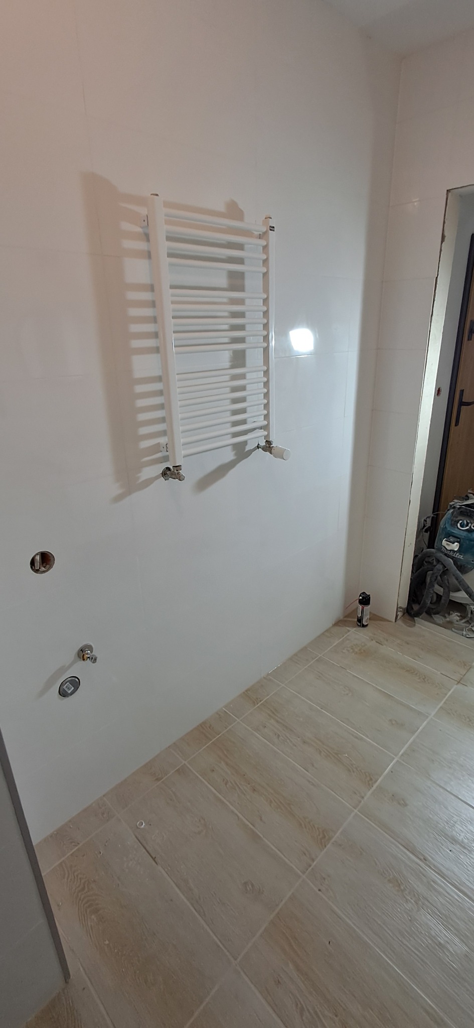 Biała łazienka w trakcie remontu: zamontowany grzejnik drabinkowy, białe płytki ścienne, drewnopodobne płytki podłogowe, widoczne przyłącza wodne i narzędzia w tle.