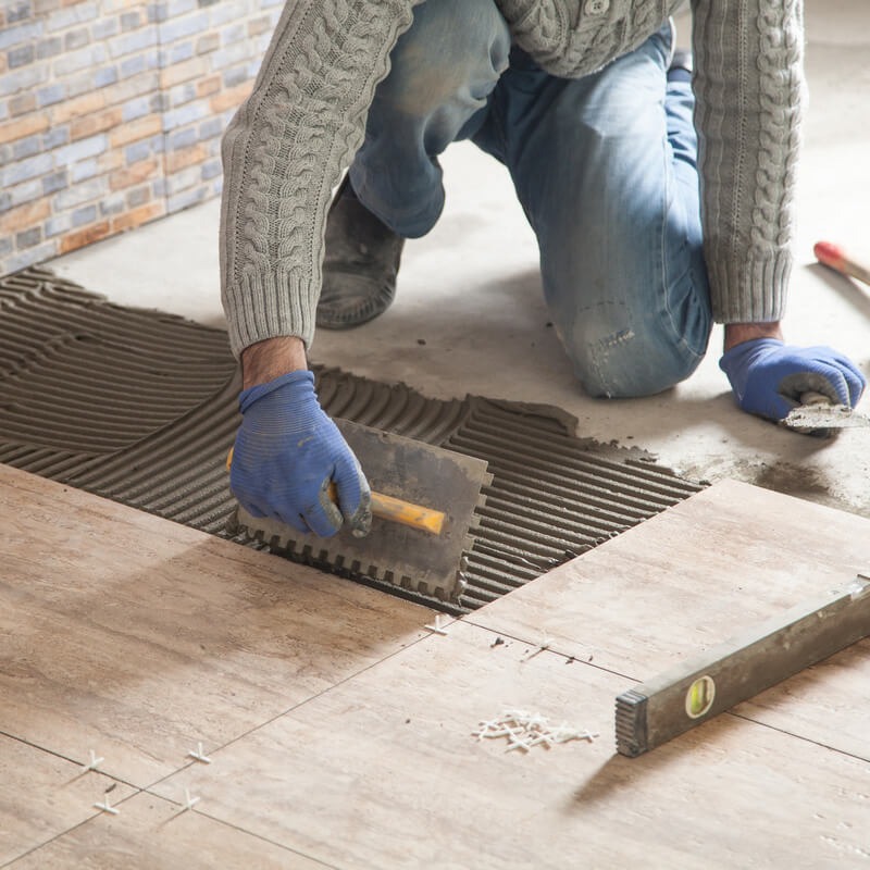 Układanie płytek podłogowych w jodełkę, z użyciem szpachli z ząbkami i poziomicy, widoczne krzyżyki dystansowe i fragment wykończonej ściany z dekoracyjnej cegły.