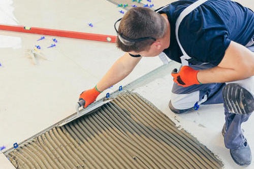 Pracownik w pomarańczowych rękawicach rozprowadza klej na płytce podłogowej za pomocą pacy z ząbkami, poziomica leży obok.