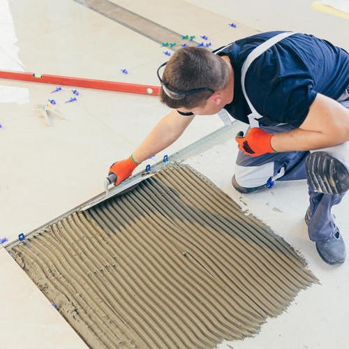 Pracownik w pomarańczowych rękawicach rozprowadza klej grzebieniem na podłodze pod płytki, używając poziomicy jako prowadnicy, widoczne niebieskie krzyżyki dystansowe.