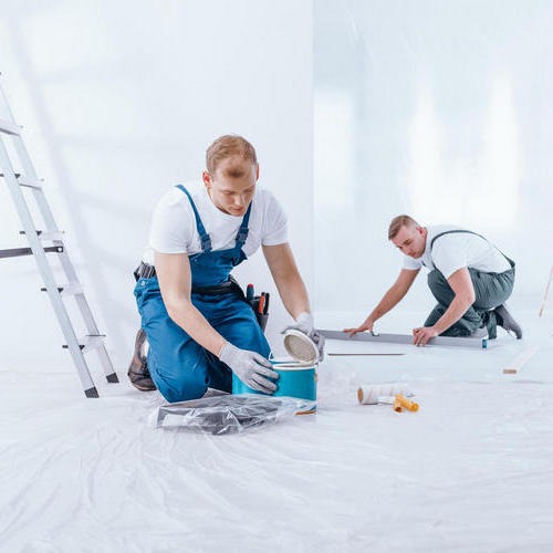 Dwóch mężczyzn w kombinezonach roboczych przygotowuje się do malowania ścian w pomieszczeniu zabezpieczonym folią ochronną; jeden otwiera puszkę z farbą, drugi używa poziomicy do wyrównania ściany.