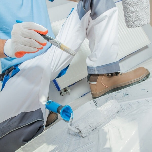 Pracownik w Łodzi maluje białą farbą podłogę wałkiem i pędzlem, widoczny kaloryfer zabezpieczony folią malarską.