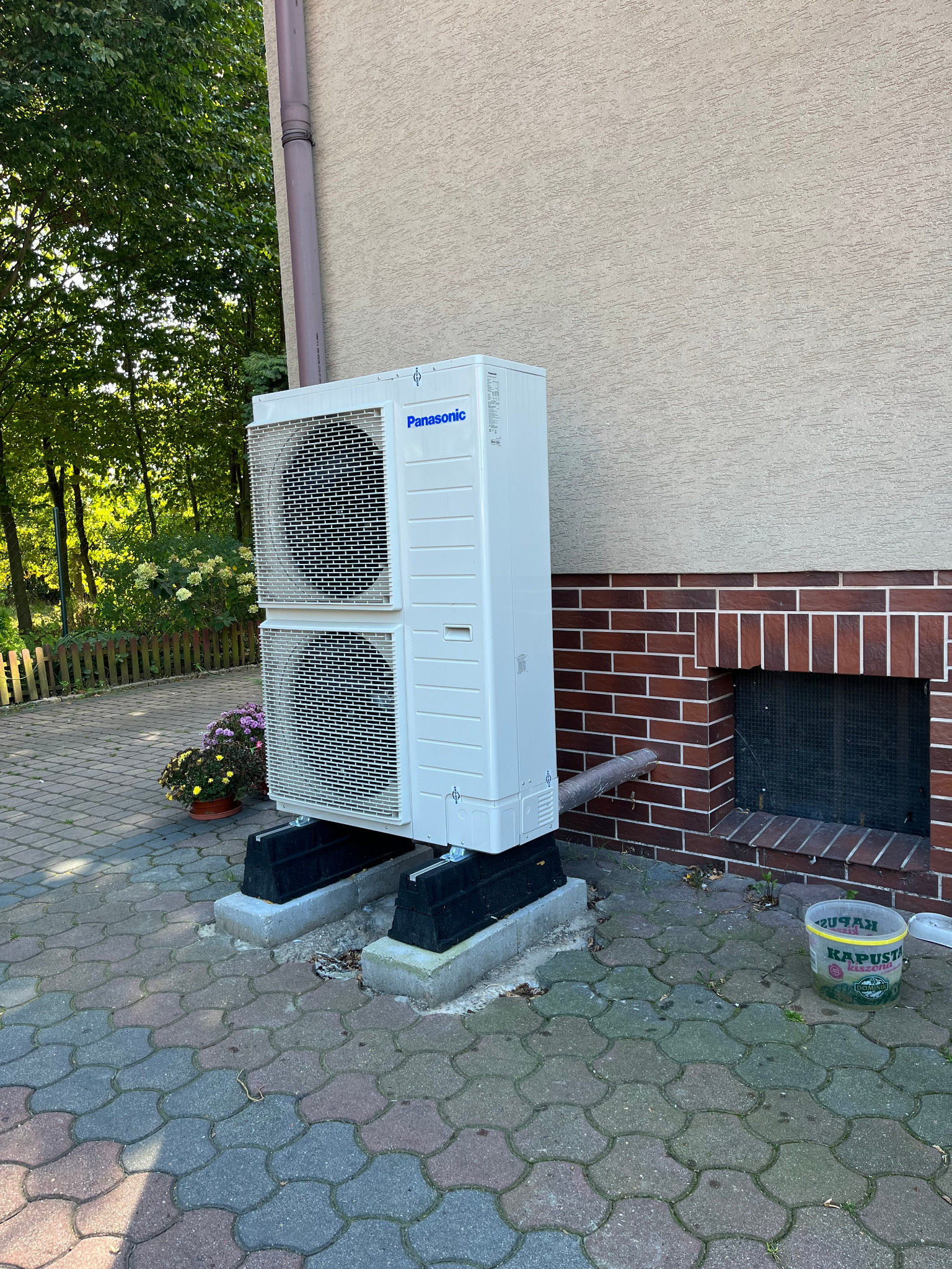 Biała jednostka zewnętrzna pompy ciepła Panasonic, umieszczona na betonowych podstawach przy ścianie budynku z ceglaną podmurówką, obok wiadro z napisem 'Kapusta Kiszona'.