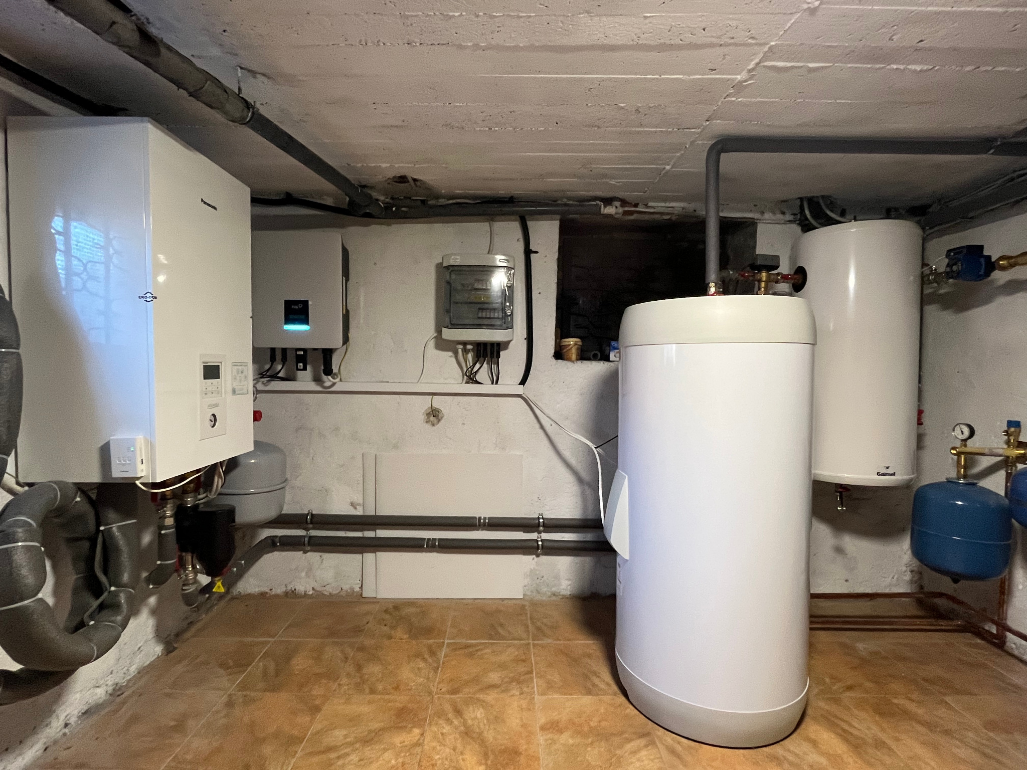 Instalacja w piwnicy z pompą ciepła Panasonic, zbiornikami buforowym i C.W.U., oraz rozdzielnicą elektryczną, widoczne rury i podłączenia.