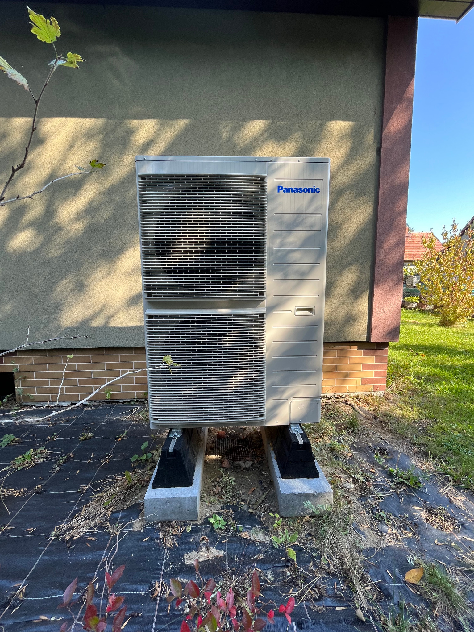 Zewnętrzna jednostka klimatyzacyjna Panasonic umieszczona na betonowych podstawach przy ścianie budynku z ceglaną podmurówką, widoczna trawa i roślinność.