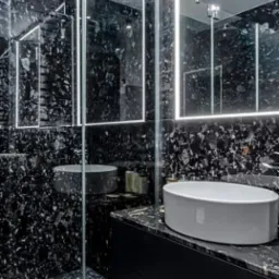 Nowoczesna łazienka z czarnymi ścianami i podłogą z efektem lastryko, biała owalna umywalka nablatowa i podświetlane lustro.