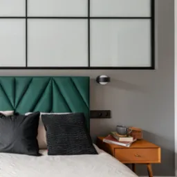 Sypialnia w odcieniach szarości z łóżkiem z zielonym zagłówkiem, czarnymi poduszkami, białą pościelą i drewnianym stolikiem nocnym z książkami i filiżanką.