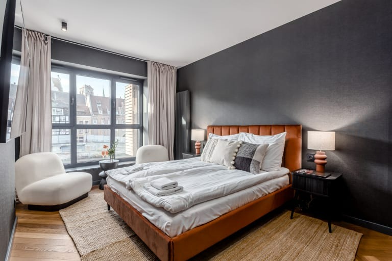 Nowoczesna sypialnia z łóżkiem z pomarańczową ramą, szarą ścianą i widokiem na miasto przez duże okno, uzupełniona o kremowe fotele i dywan z juty.