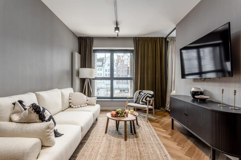 Nowoczesny salon z białą sofą, drewnianym parkietem w jodełkę, ciemną komodą pod telewizorem i widokiem na miasto za oknem.