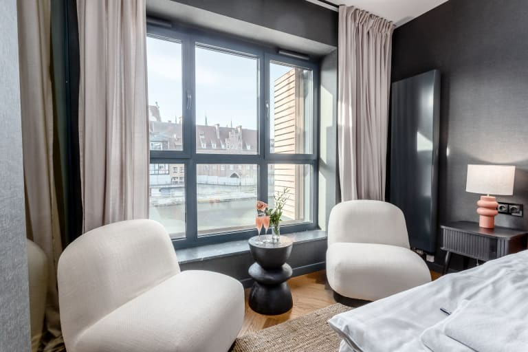 Nowoczesna sypialnia z dużym oknem z widokiem na rzekę, dwa białe fotele, czarny stolik z kwiatami i kieliszkami, łóżko z białą pościelą, lampka nocna w kolorze koralowym.