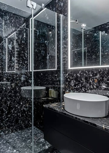 Nowoczesna łazienka z czarnymi ścianami i podłogą z efektem lastryko, biała owalna umywalka nablatowa i podświetlane lustro.