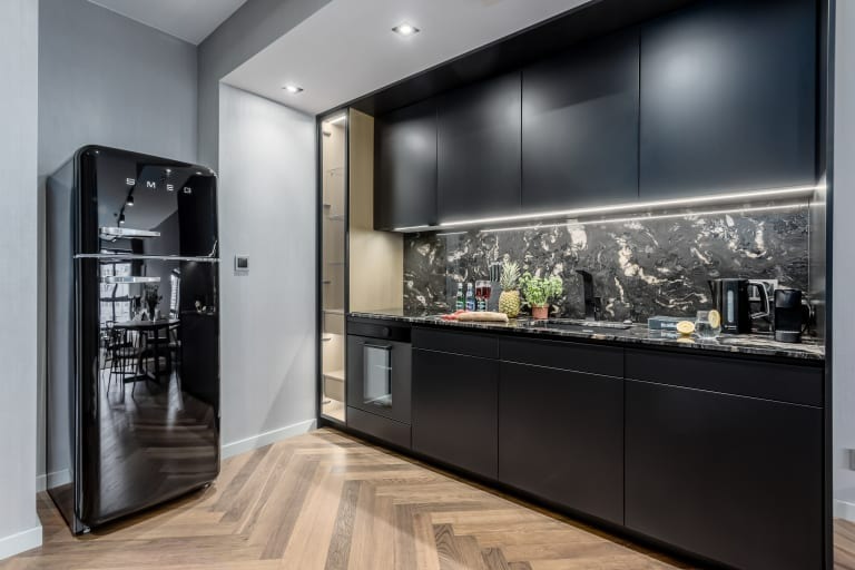 Nowoczesna kuchnia w ciemnych barwach z czarnymi szafkami, blatem z marmuru i lodówką Smeg, oświetlona ledami i światłem punktowym, z podłogą ułożoną w jodełkę.