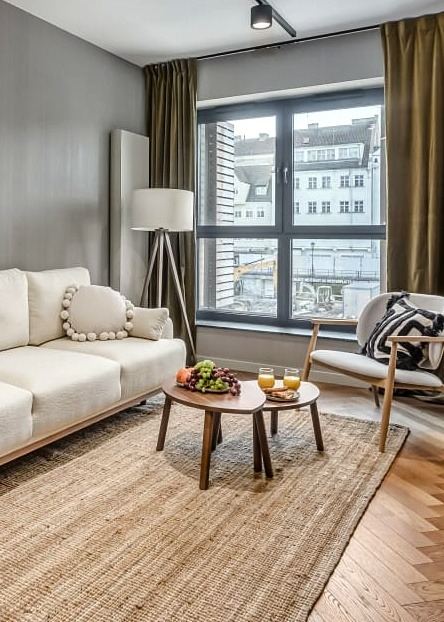 Jasny salon z beżową sofą, drewnianym stolikiem kawowym z owocami i sokiem, oraz fotelem z poduszką w paski, dywanem z juty i dużym oknem z widokiem na miasto.