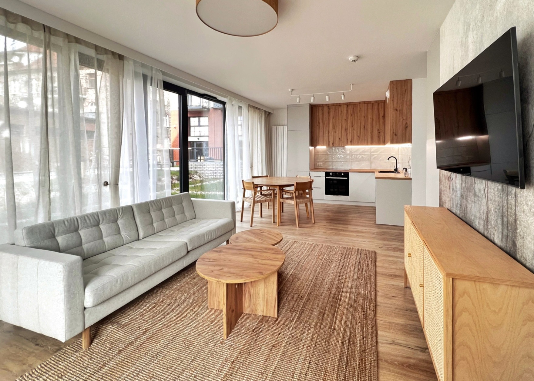 Jasne, przestronne wnętrze mieszkania z otwartą kuchnią, szarą sofą z poduszkami, drewnianymi stolikami kawowymi, dywanem z juty i telewizorem na ścianie z efektem betonu.