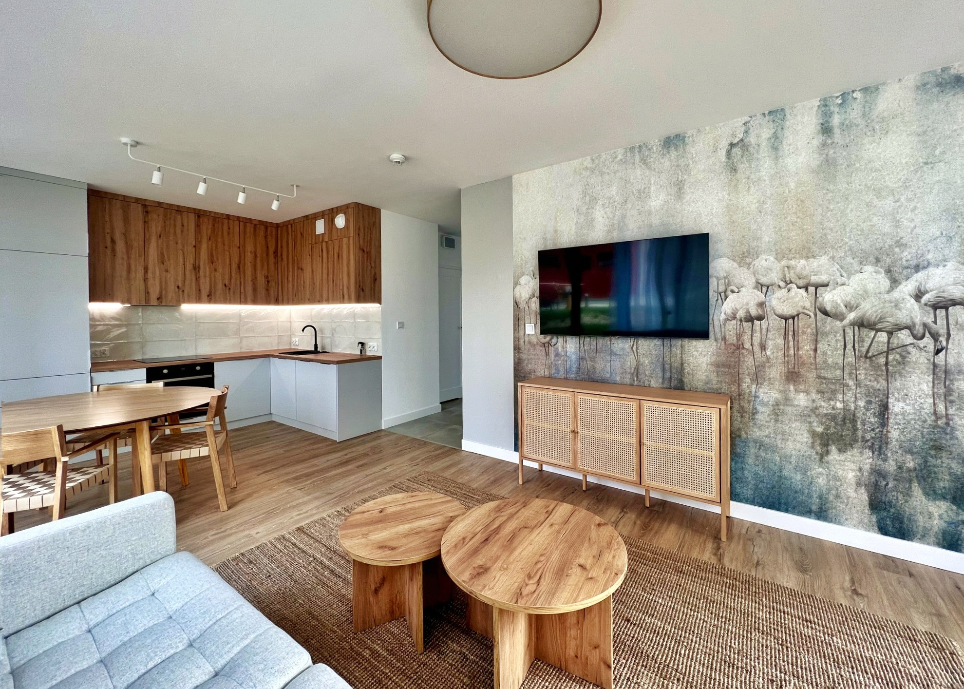 Nowoczesne wnętrze salonu z aneksem kuchennym, z drewnianymi meblami, tapetą z motywem flamingów, okrągłymi stolikami kawowymi i szarą sofą.