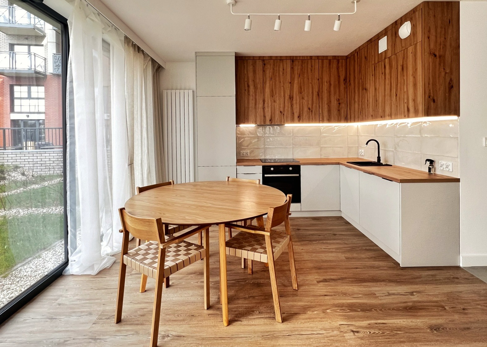 Jasna, nowoczesna kuchnia z drewnianymi szafkami górnymi i białymi dolnymi, okrągły drewniany stół z czterema krzesłami z plecionymi siedziskami, duże okno z widokiem na budynek z czerwonej cegły.