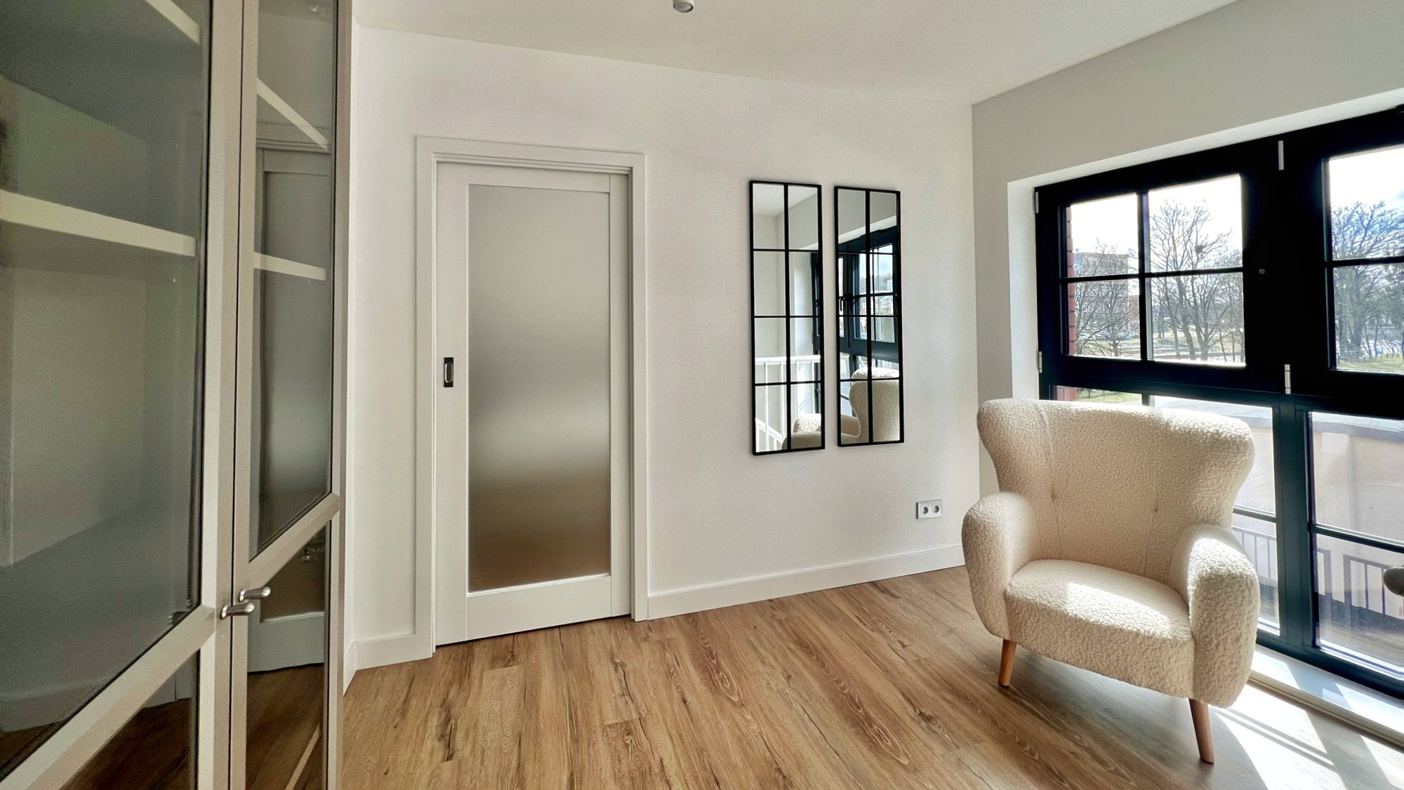 Jasne wnętrze w Gdyni z drewnianą podłogą, kremowym fotelem typu uszak przy czarnym oknie, szklaną szafą i lustrami w czarnych ramach na białej ścianie.