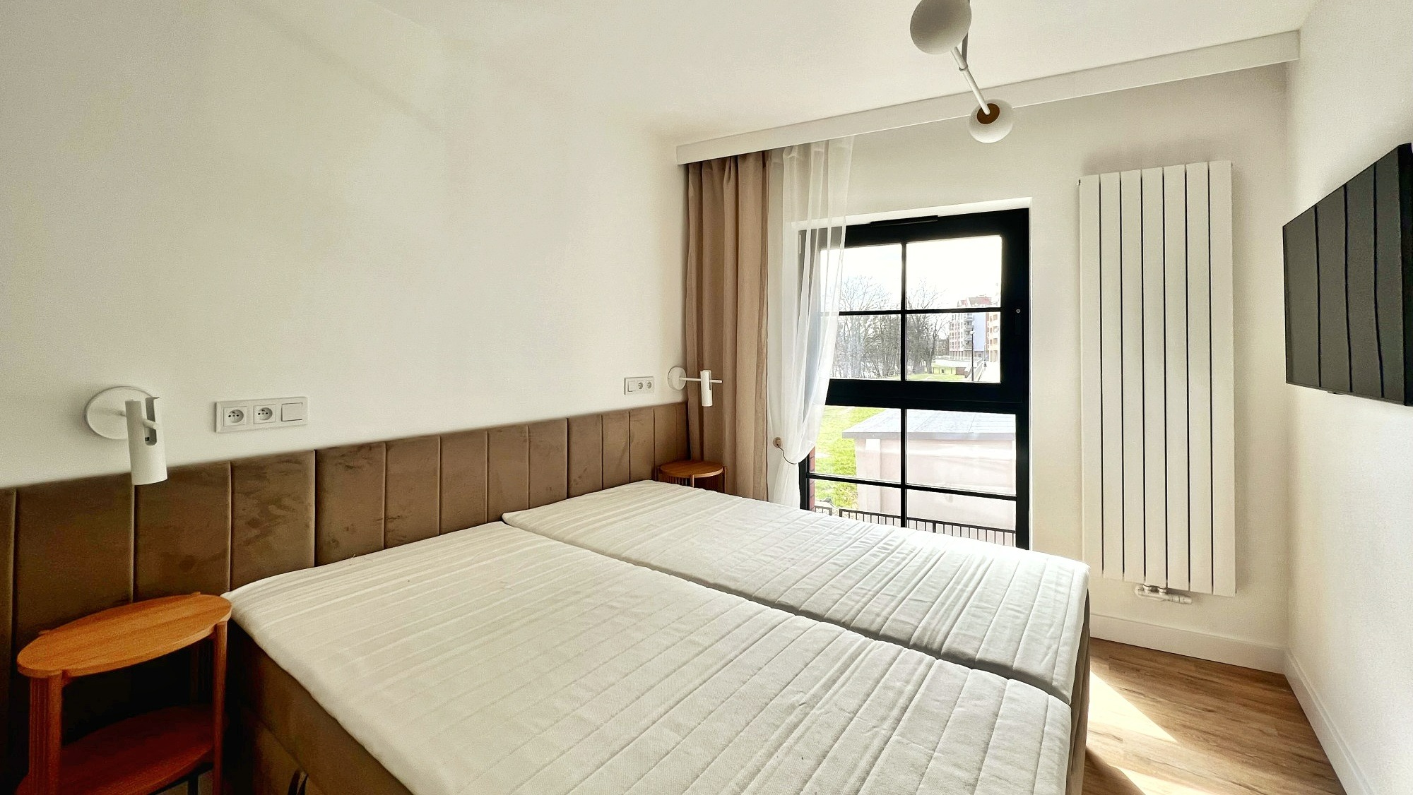 Jasna sypialnia z dwoma łóżkami z brązowym wezgłowiem, stolikiem nocnym, oknem z czarną ramą i grzejnikiem panelowym.