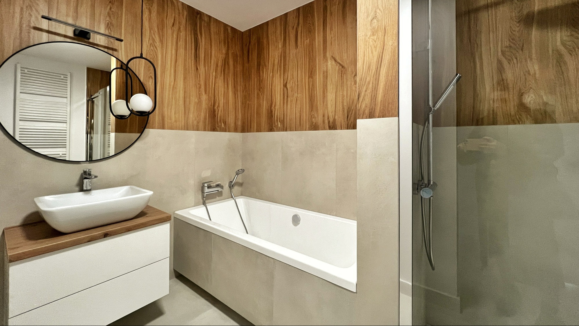Nowoczesna łazienka z drewnianymi akcentami: okrągłe lustro z podświetleniem, umywalka nablatowa na drewnianym blacie, biała wanna i kabina prysznicowa z chromowaną armaturą, ściany wykończone...
