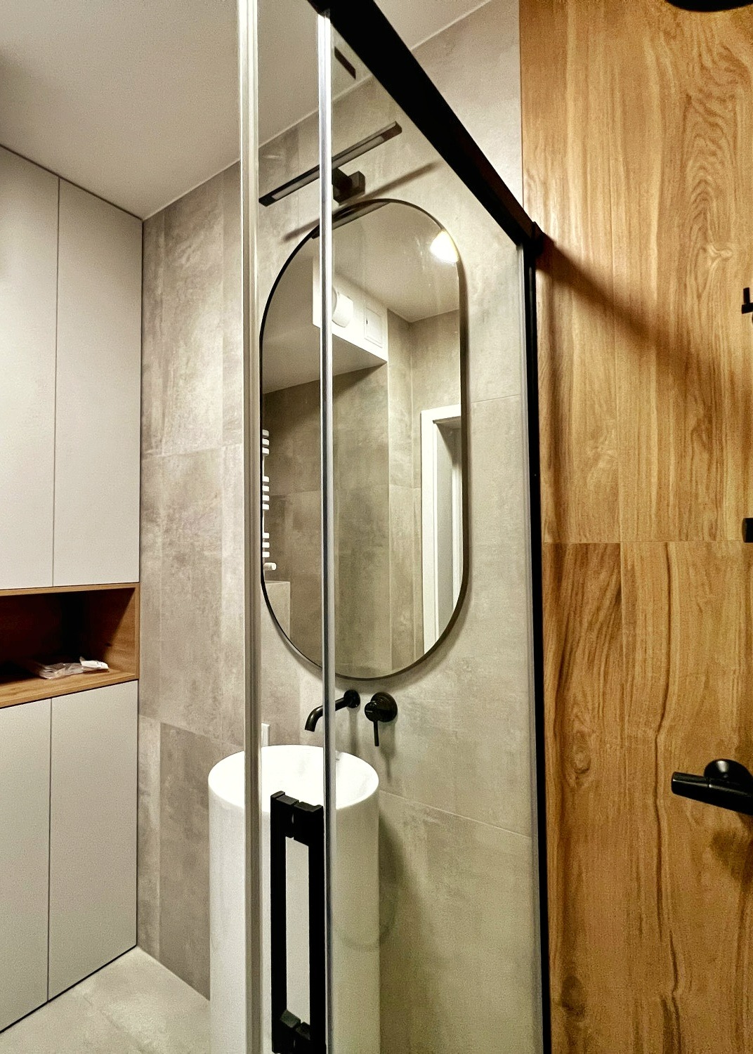 Nowoczesna łazienka z okrągłym lustrem w czarnej ramie, cylindryczną białą umywalką i drewnianymi drzwiami, widok zza szklanej kabiny prysznicowej.