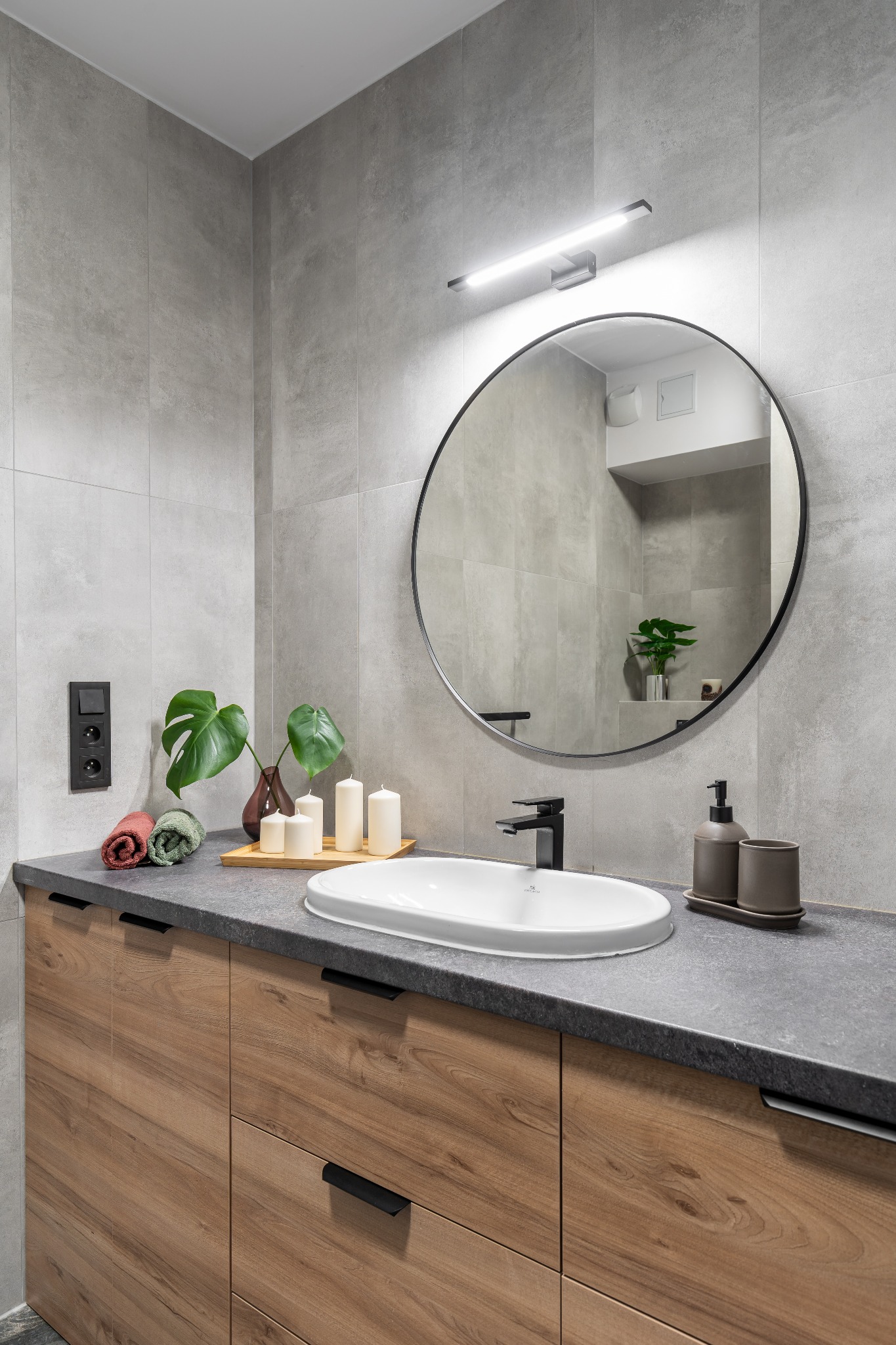 Nowoczesna łazienka z szarymi płytkami, drewnianą szafką pod umywalką z czarnymi uchwytami, okrągłym lustrem z podświetleniem LED i dekoracjami w postaci świec, ręczników i rośliny w wazonie.