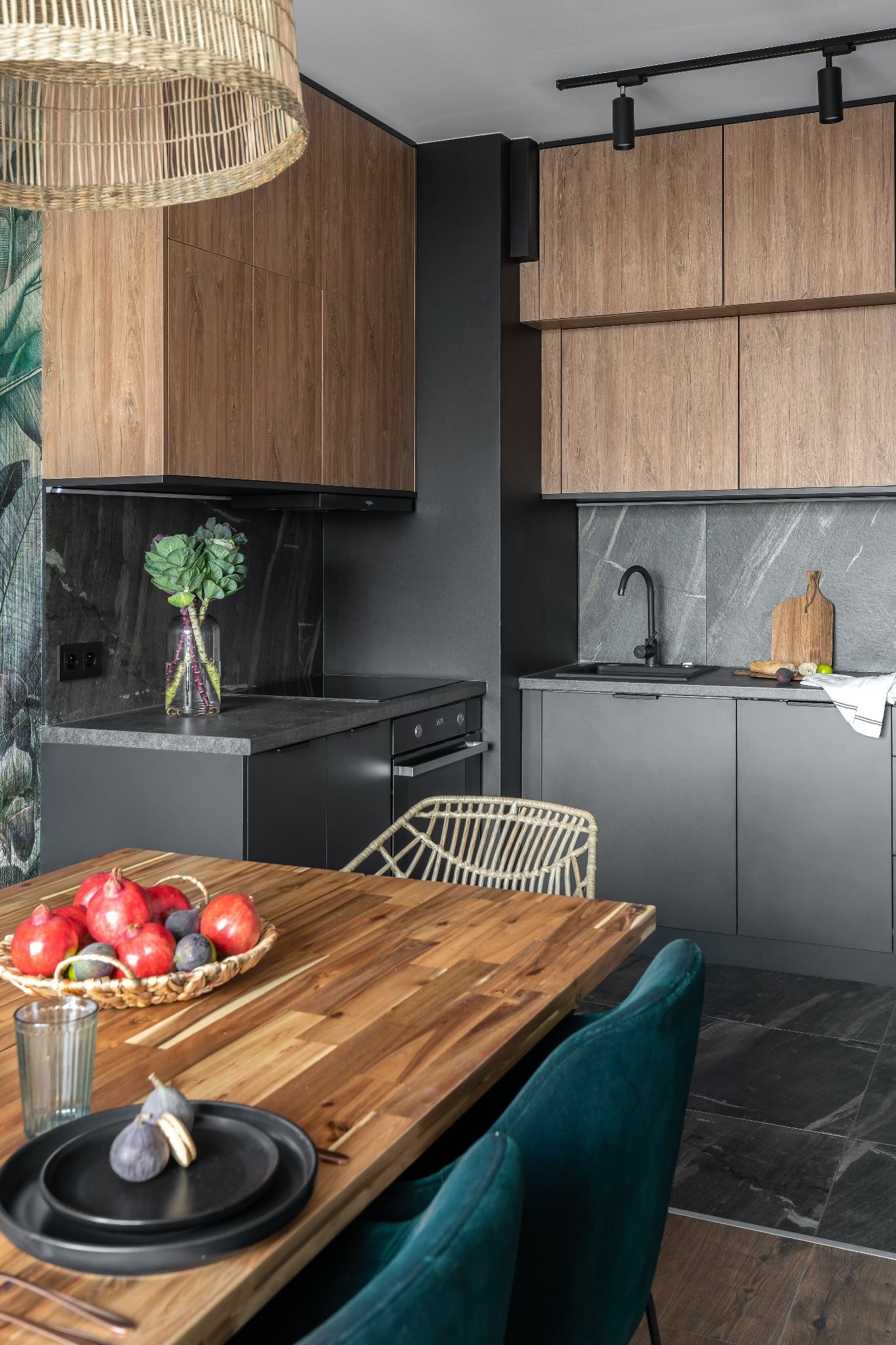 Nowoczesna kuchnia z szarymi szafkami i drewnianymi frontami, drewniany stół z owocami i ciemnozielonymi krzesłami, lampa z wikliny