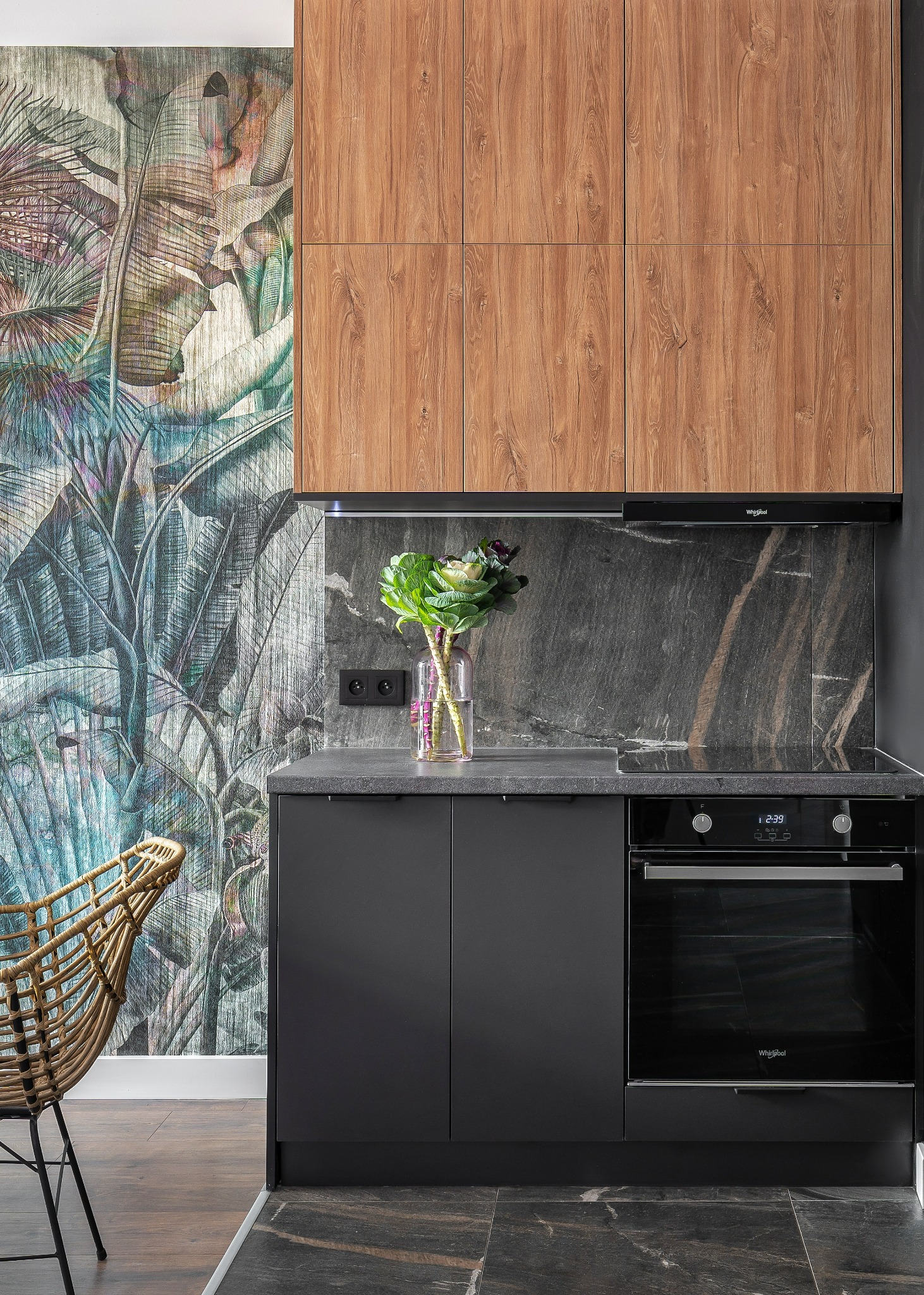 Nowoczesna kuchnia z ciemnymi szafkami, drewnianymi górnymi szafkami i tapetą w tropikalne liście. Na blacie stoi wazon z kwiatami, a obok rattanowe krzesło.