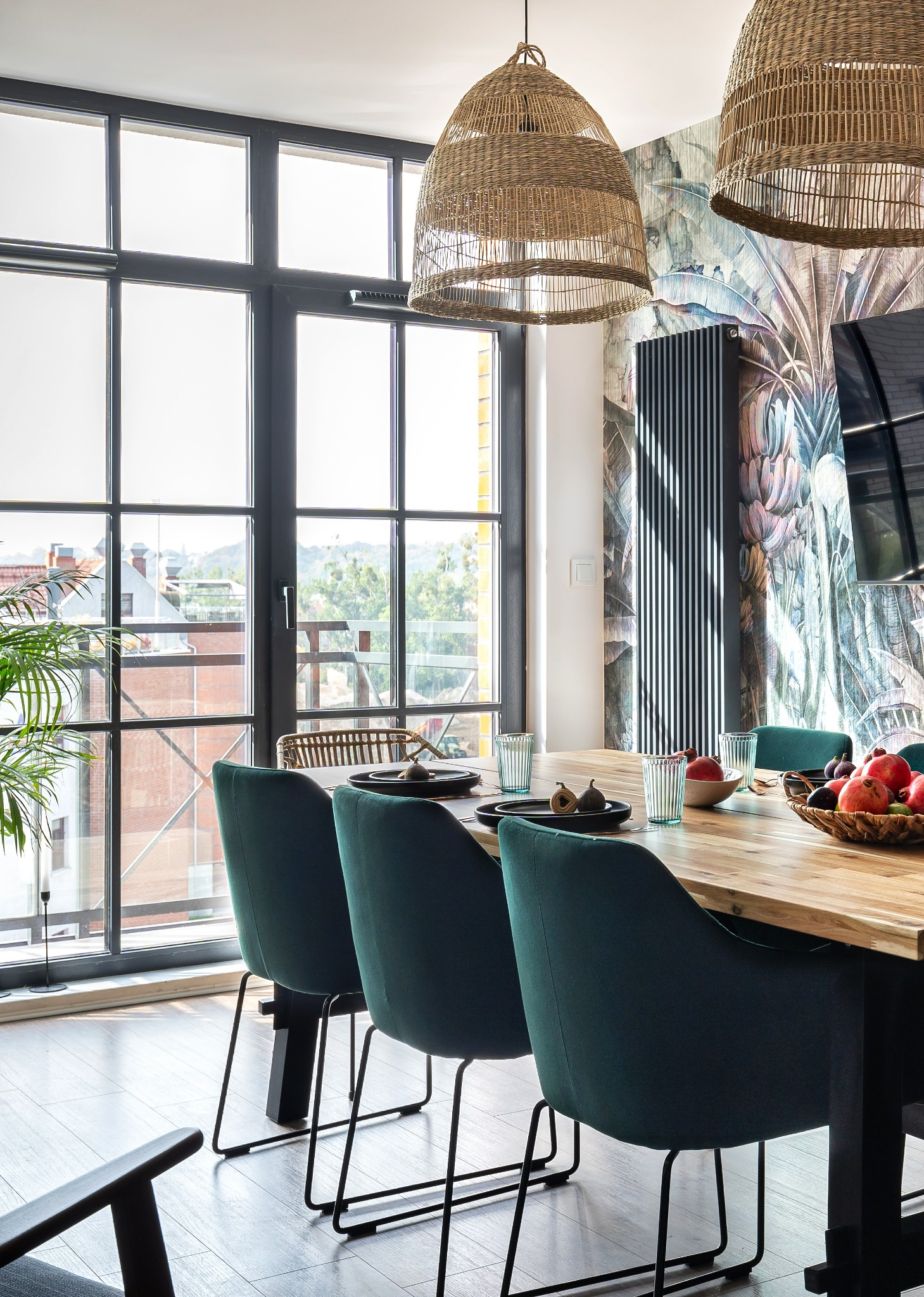 Jadalnia z drewnianym stołem, zielonymi krzesłami i wiklinowymi lampami na tle okna z widokiem na miasto i tapety w motywy roślinne.