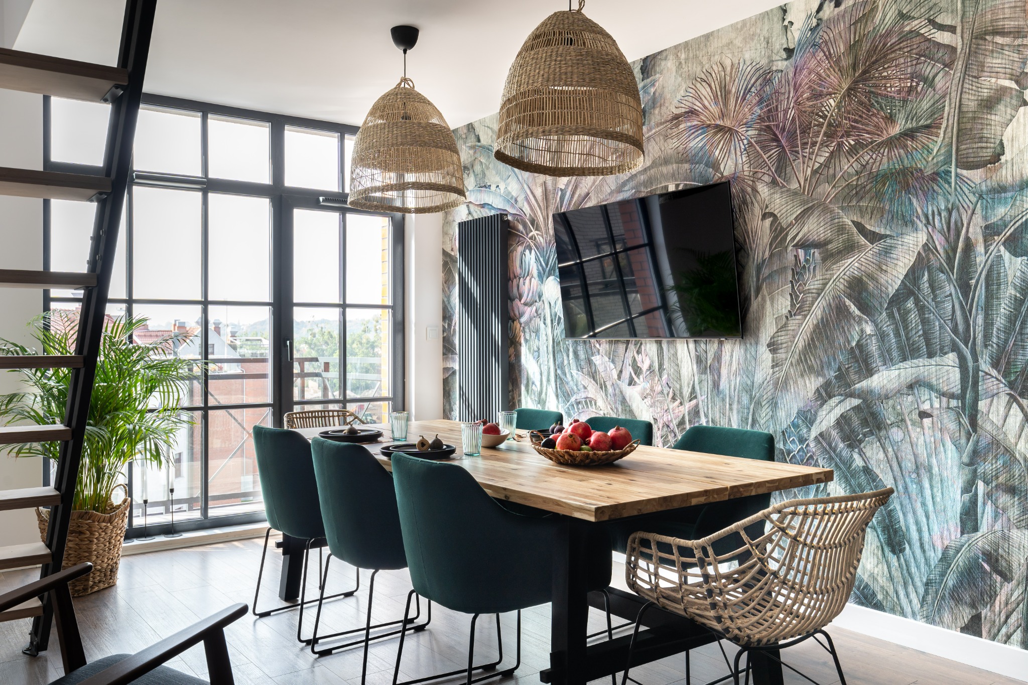 Jadalnia w mieszkaniu z drewnianym stołem, krzesłami tapicerowanymi i wiklinowymi, lampami z rattanu i tapetą w tropikalne liście.