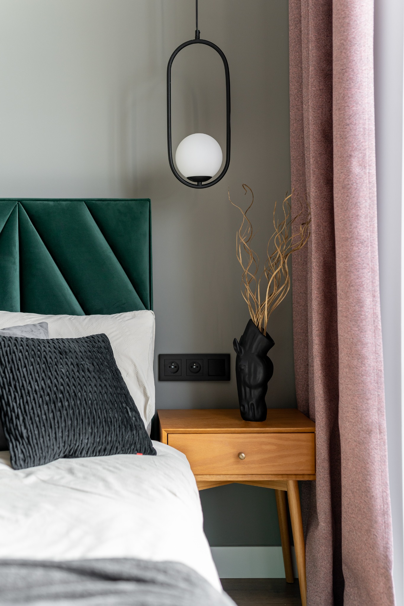 Sypialnia z zielonym, pikowanym zagłówkiem, czarną poduszką, lampą z kulistym kloszem, drewnianą szafką nocną z czarnym wazonem w kształcie głowy konia i zasłonami w odcieniach różu.