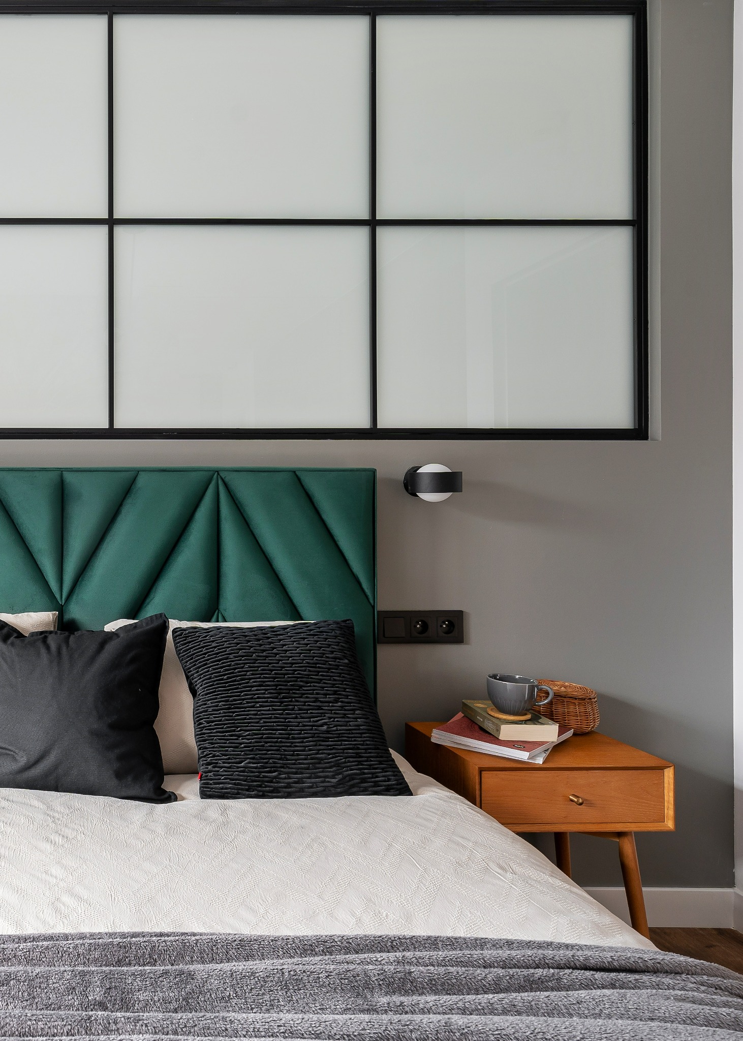 Sypialnia w odcieniach szarości z łóżkiem z zielonym zagłówkiem, czarnymi poduszkami, białą pościelą i drewnianym stolikiem nocnym z książkami i filiżanką.